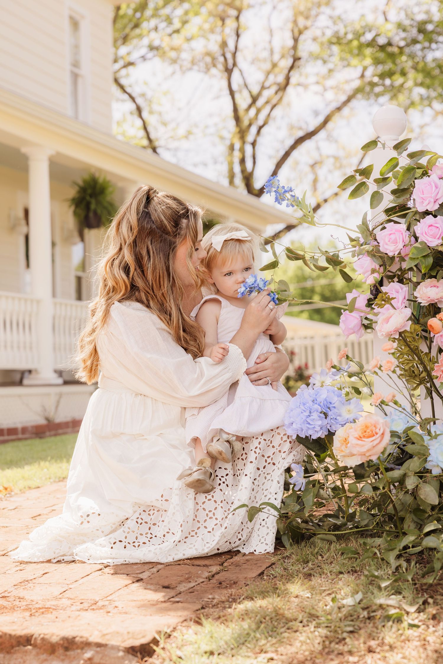 Mother in white dress kissing baby girl among colorful spring flowers outside a charming white farmhouse.