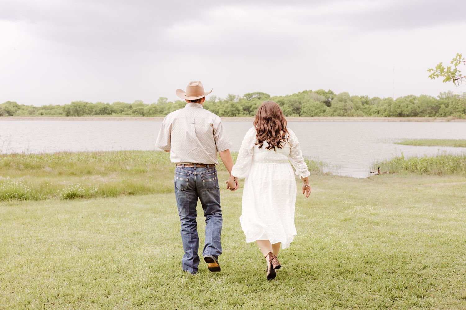 Couple holding hands walking toward a serene lake on green grass, man in hat and jeans, woman in white dress.