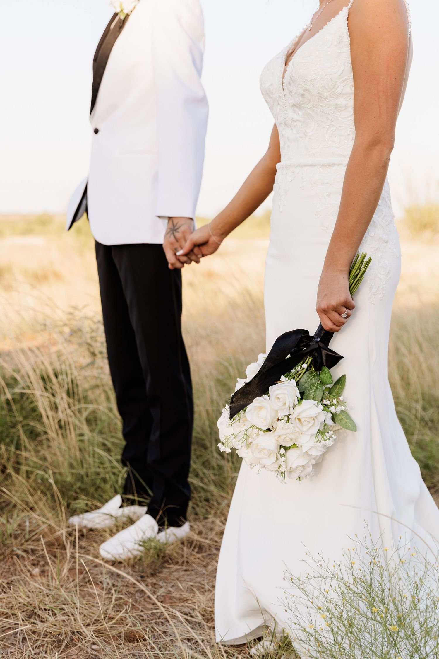 Bride holding white rose bouquet with black ribbon, holding hands with groom in white jacket and black pants outdoors.