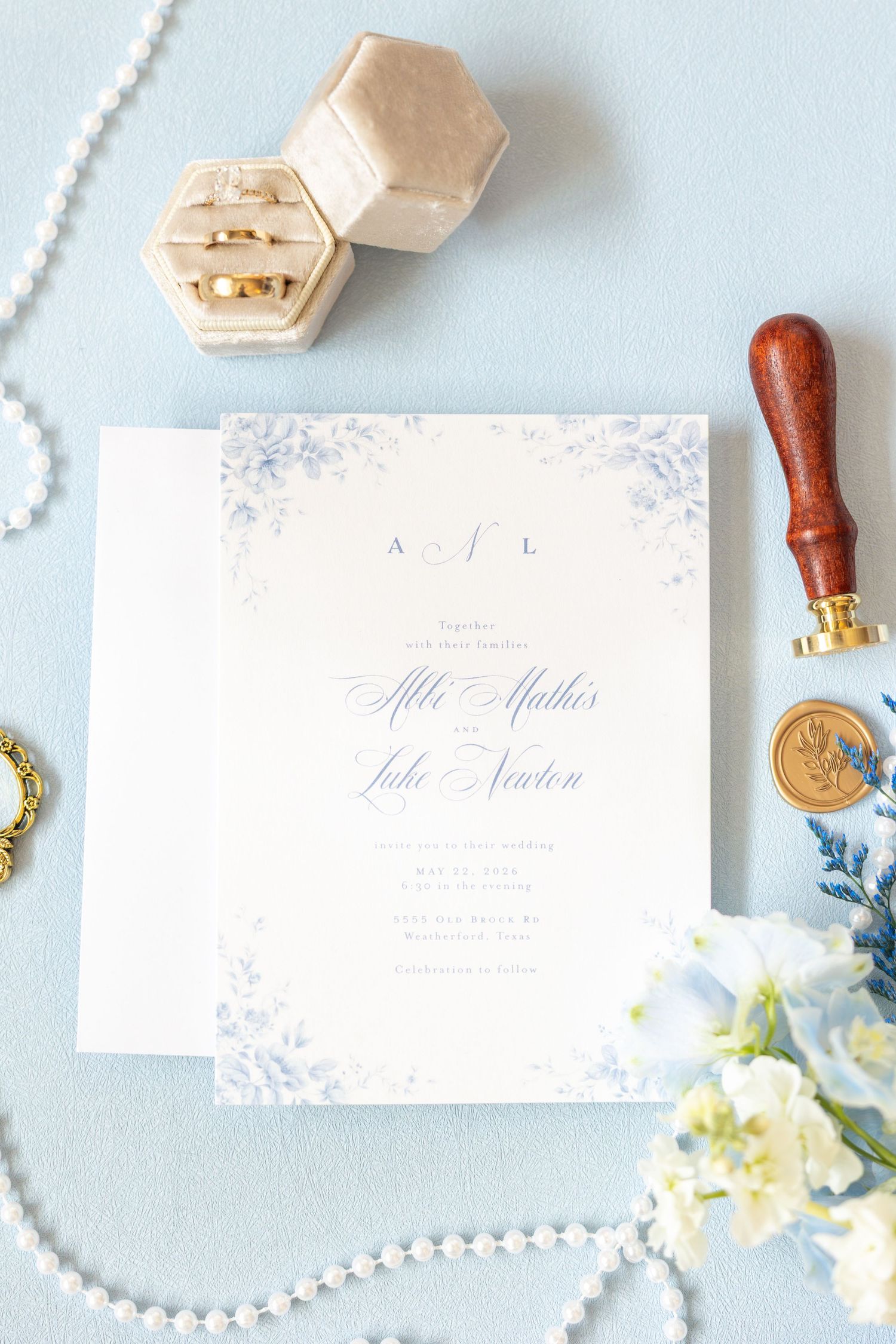 Elegant wedding invitation suite with blue floral details, gold rings in velvet box, wax seal stamp, and pearl necklace on blue background.