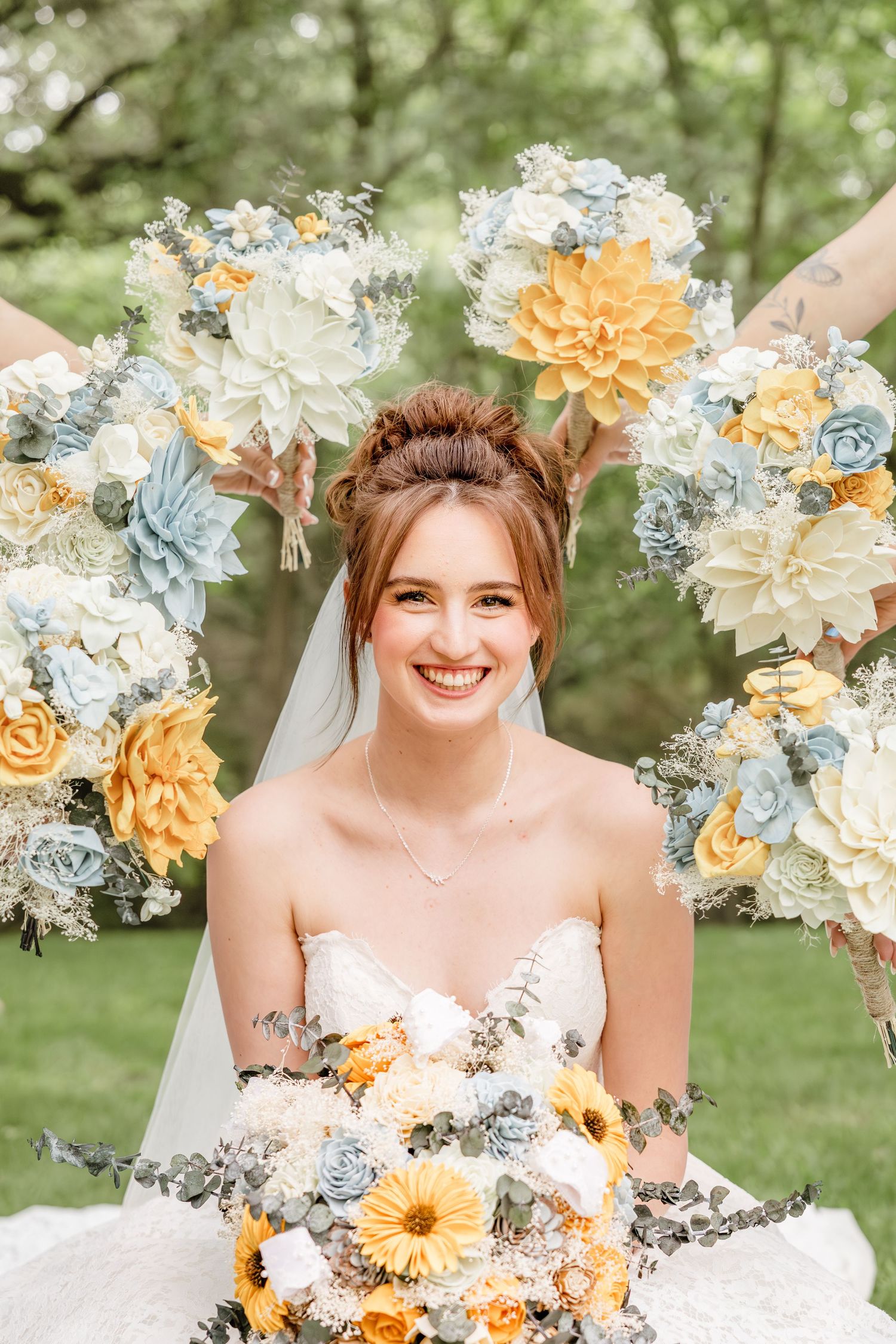 Smiling bride in strapless gown holds yellow and blue floral bouquet, surrounded by matching bridesmaid bouquets outdoors.