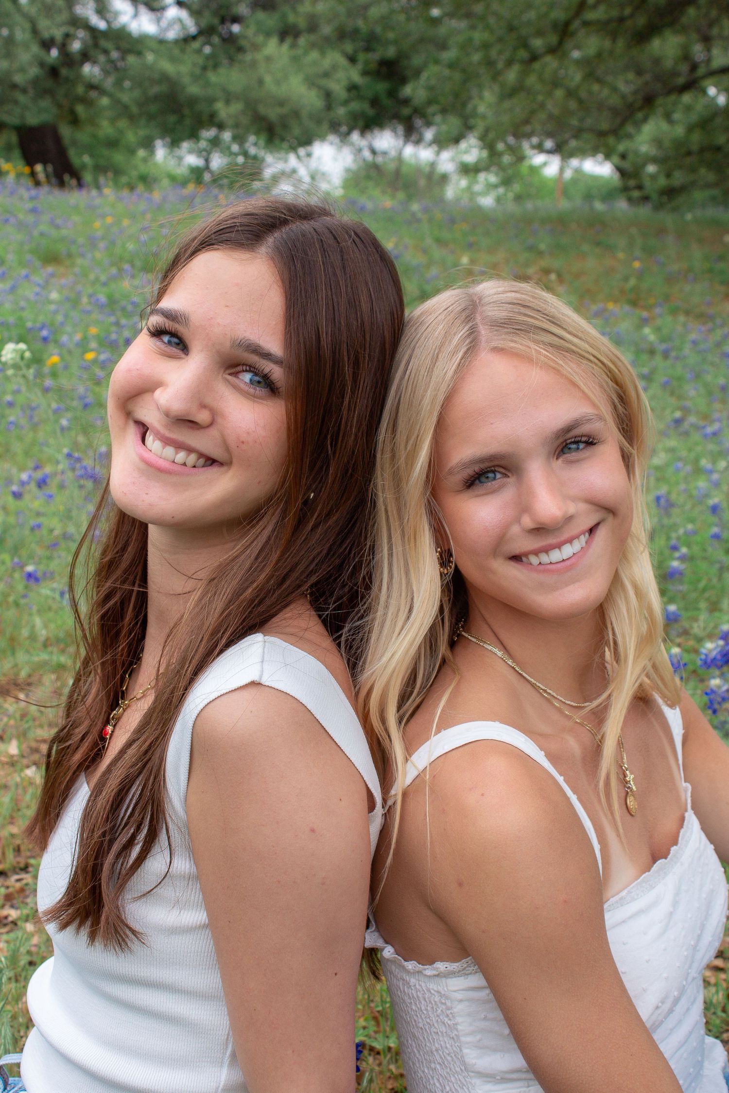 Two friends in white tops pose together outdoors in a garden setting with flowers and greenery in the background.