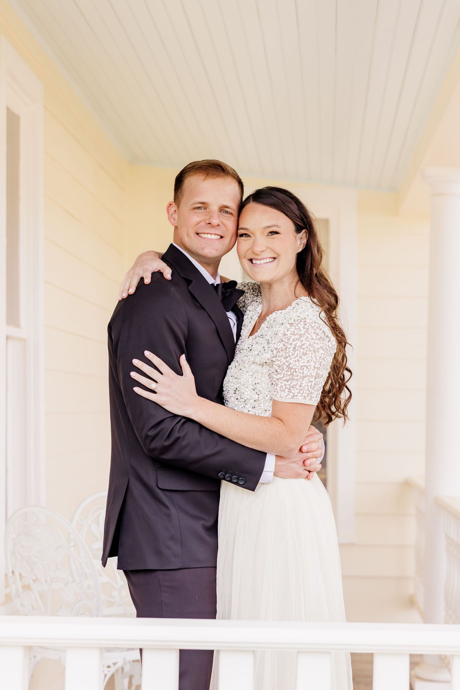 A smiling couple in wedding attire embraces on a white porch, the groom in a dark suit and bride in a white gown.