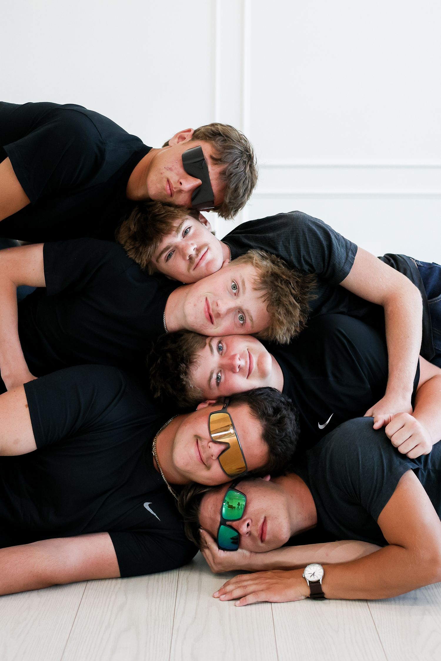 Group of friends wearing black shirts lay in a pile while wearing colorful sunglasses.