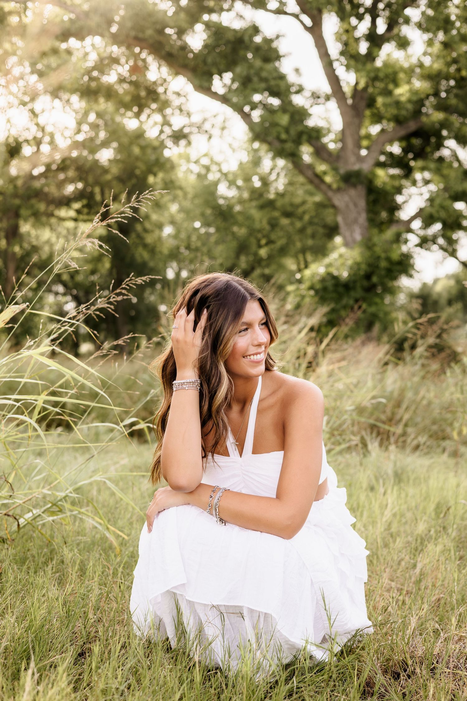 Young woman in white dress sitting in tall grass, smiling, with sunlit trees in background.
