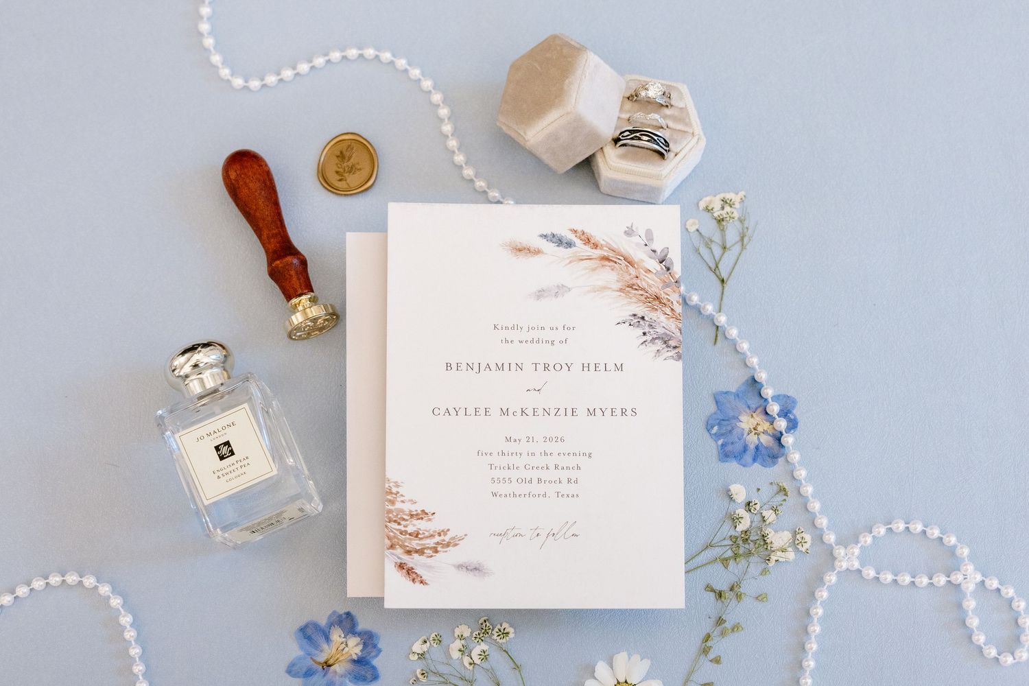 Elegant wedding invitation flatlay with perfume, wax seal, ring box, and blue florals on a soft blue background.