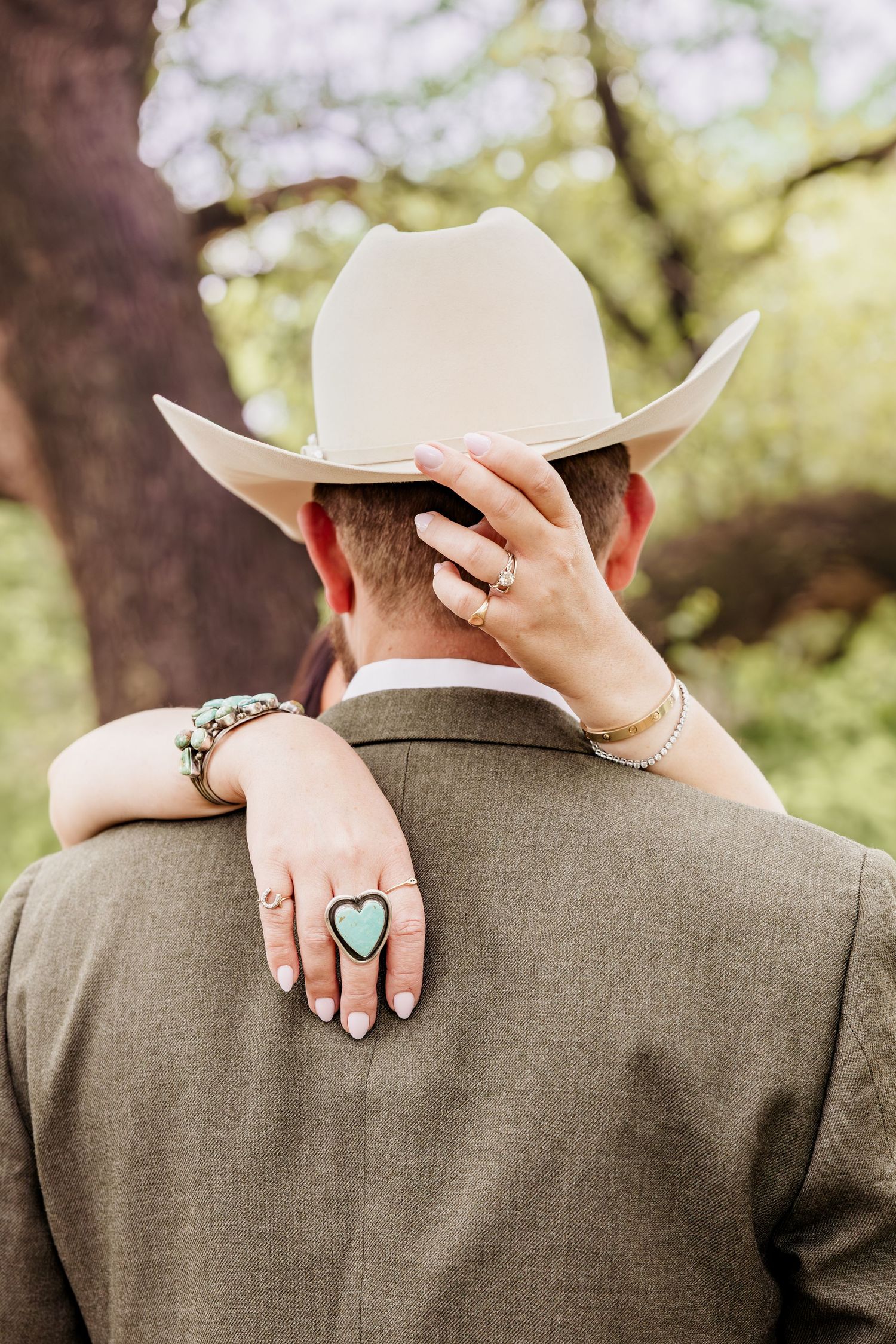 Couple in western wedding attire, woman's hand with turquoise heart ring resting on man's shoulder in tan cowboy hat.