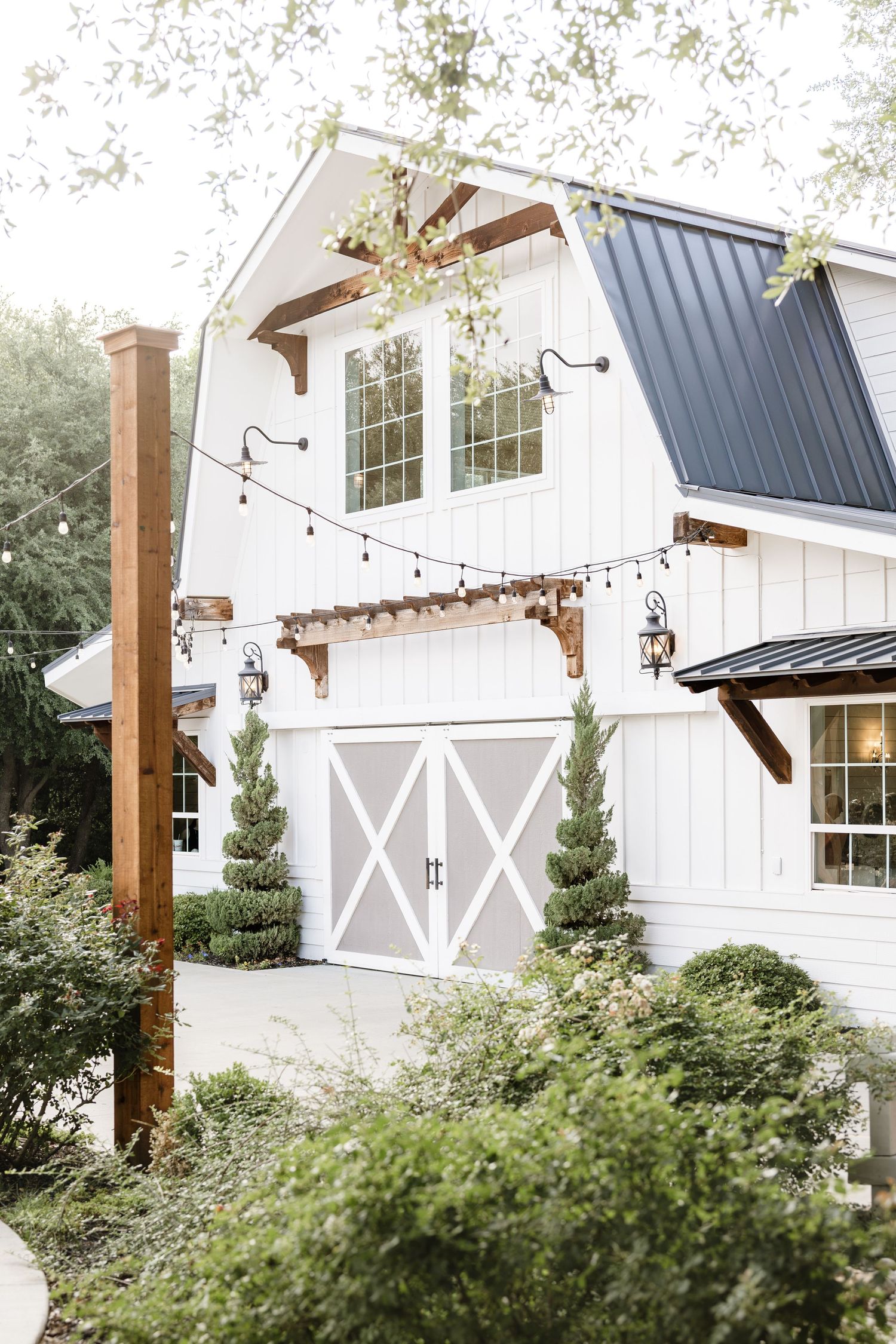 White farmhouse with black metal roof, barn-style garage doors, string lights, and lush greenery in a charming exterior.