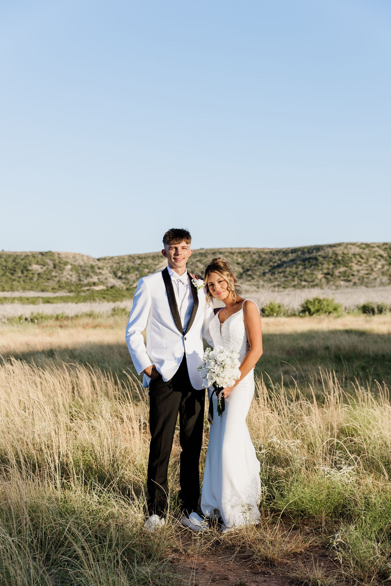 Bride and groom posing in a golden field with scenic hills, bride holding white bouquet, groom in white tuxedo jacket.