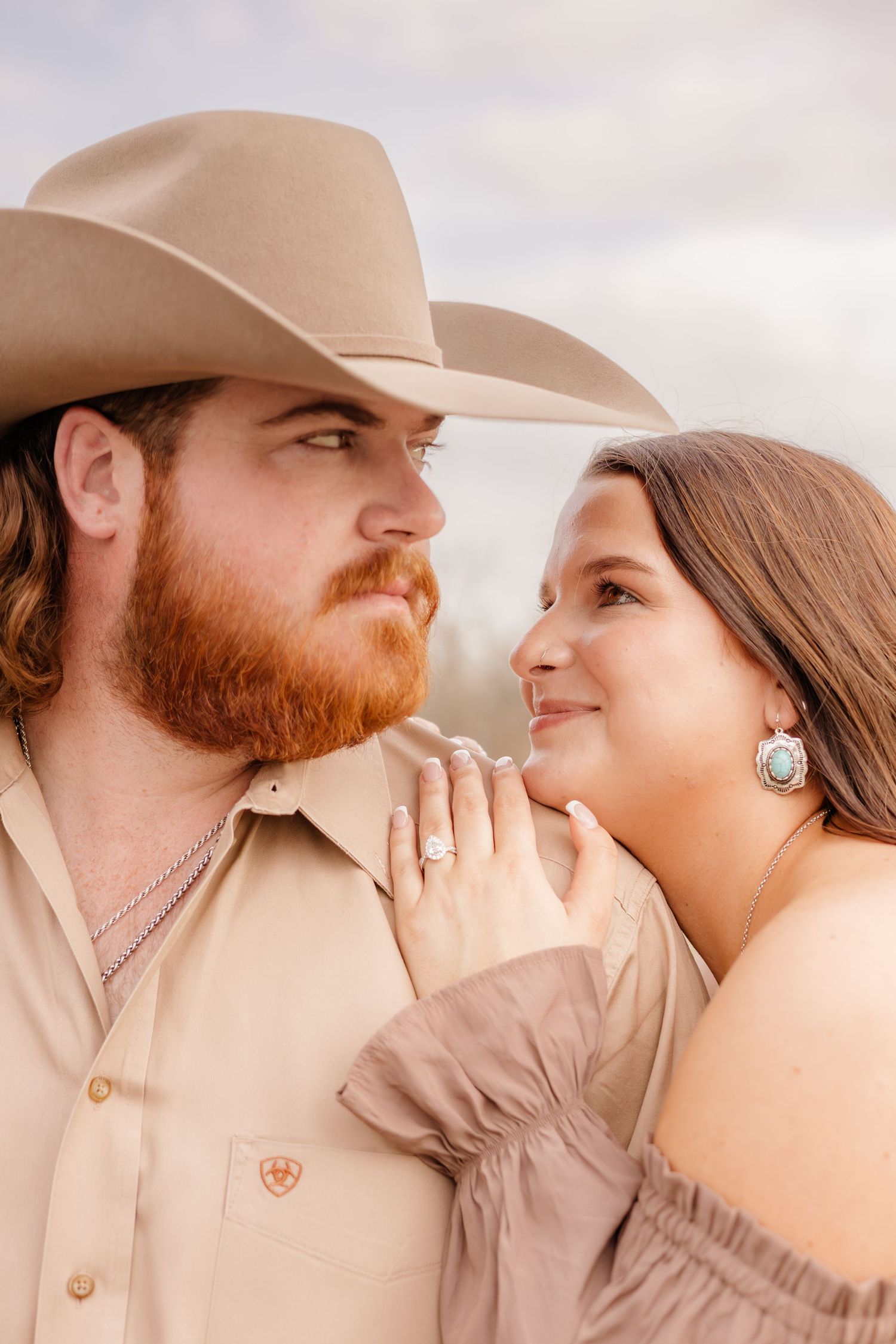 Couple in western attire sharing a romantic moment, man in cowboy hat with red beard, woman showing engagement ring.