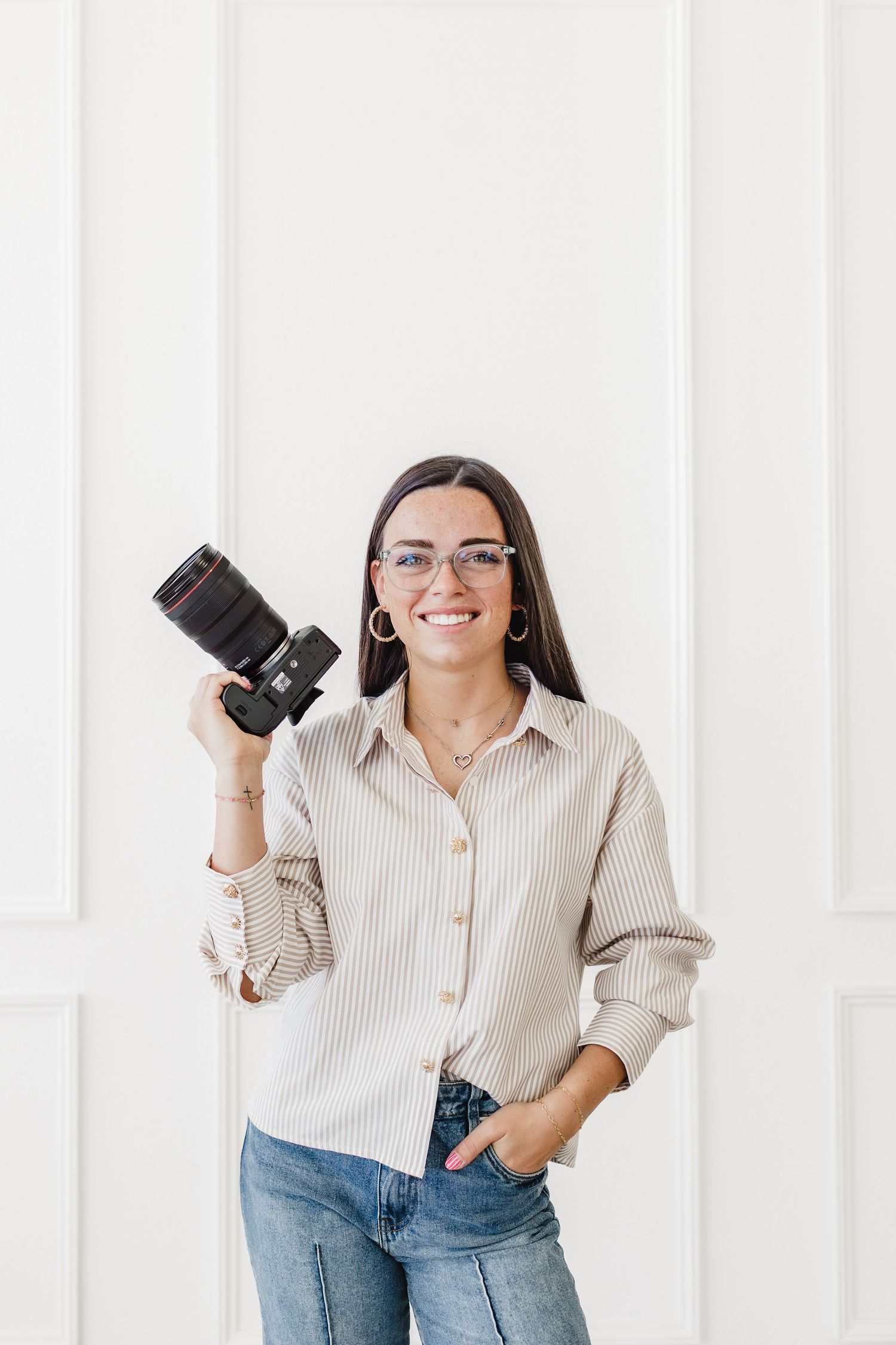 Smiling female photographer holding DSLR camera, wearing glasses and striped shirt, posing against white wall.