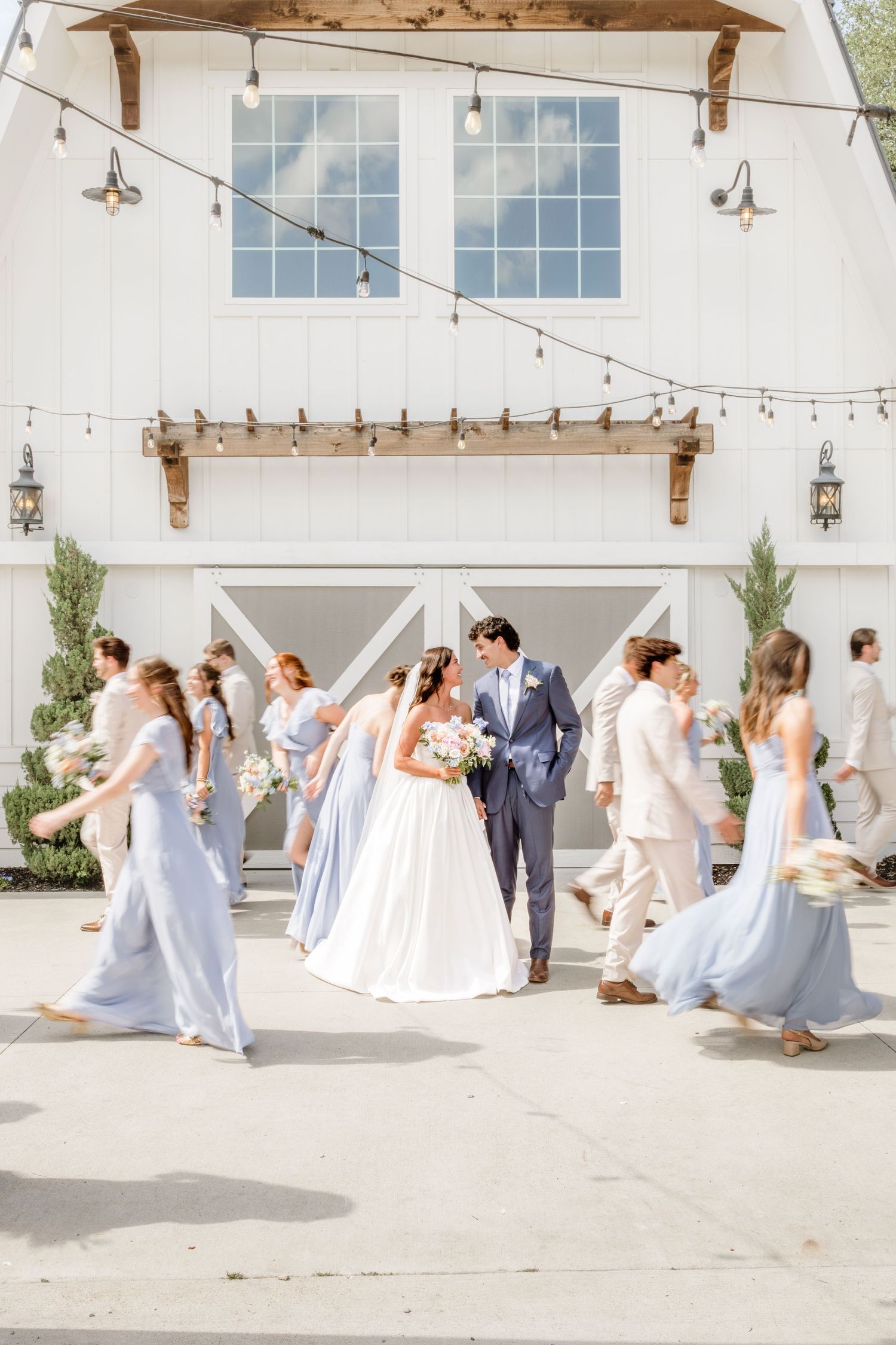 Bride and groom share a romantic moment surrounded by bridesmaids in blue gowns and groomsmen outside a white barn venue.