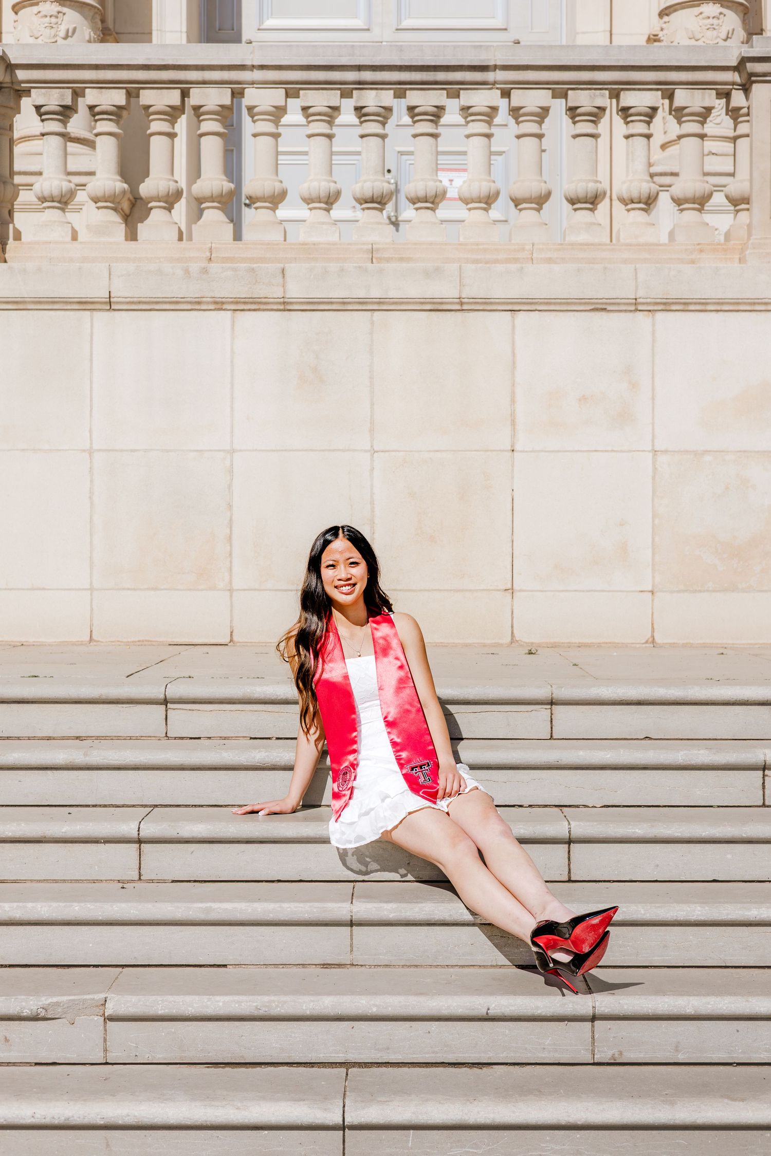 Young woman in white dress and red graduation stole sitting on stone steps of a classical building, smiling at camera.