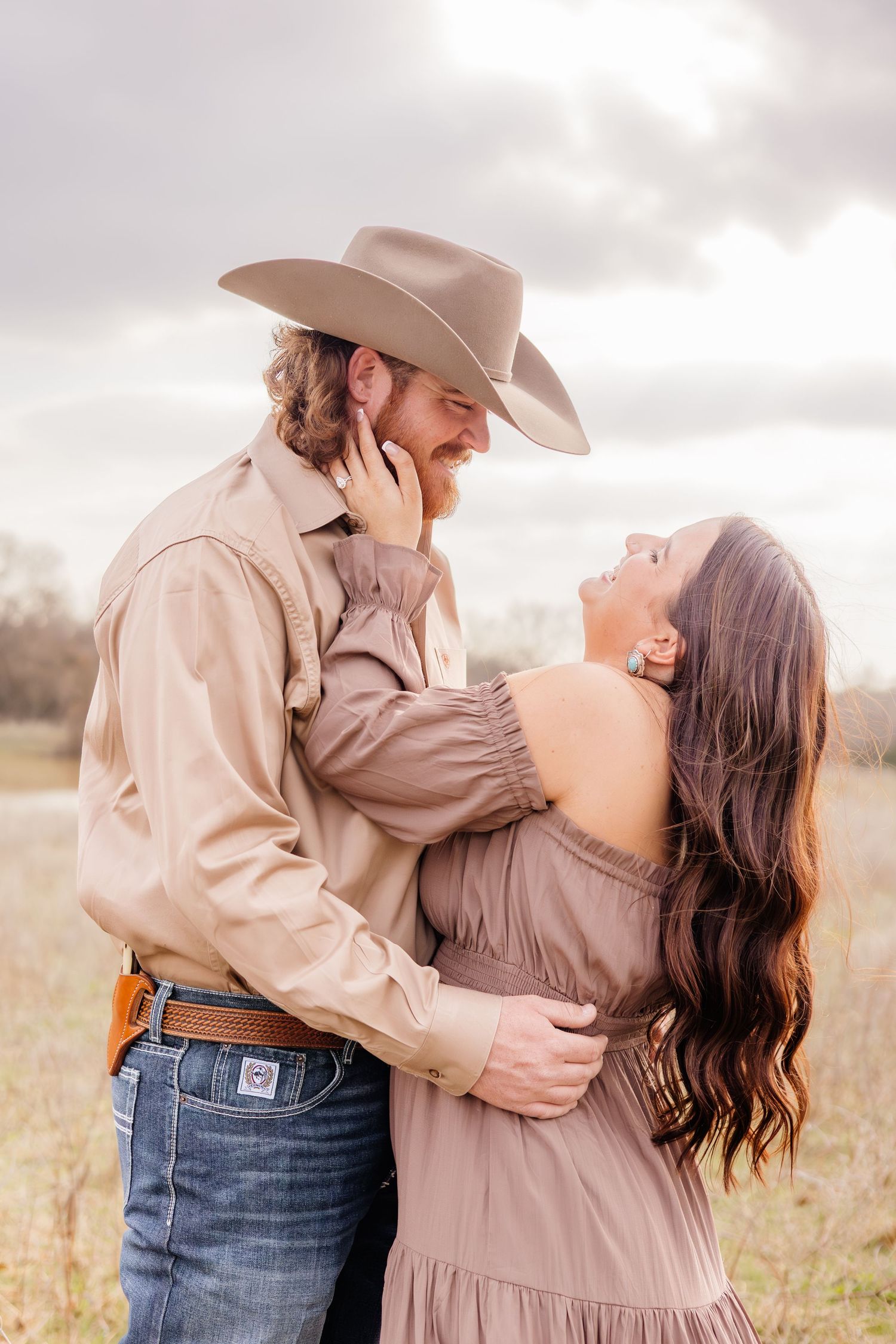 A couple in Western attire embraces in a field, the man in a cowboy hat and the woman in an off-shoulder dress.