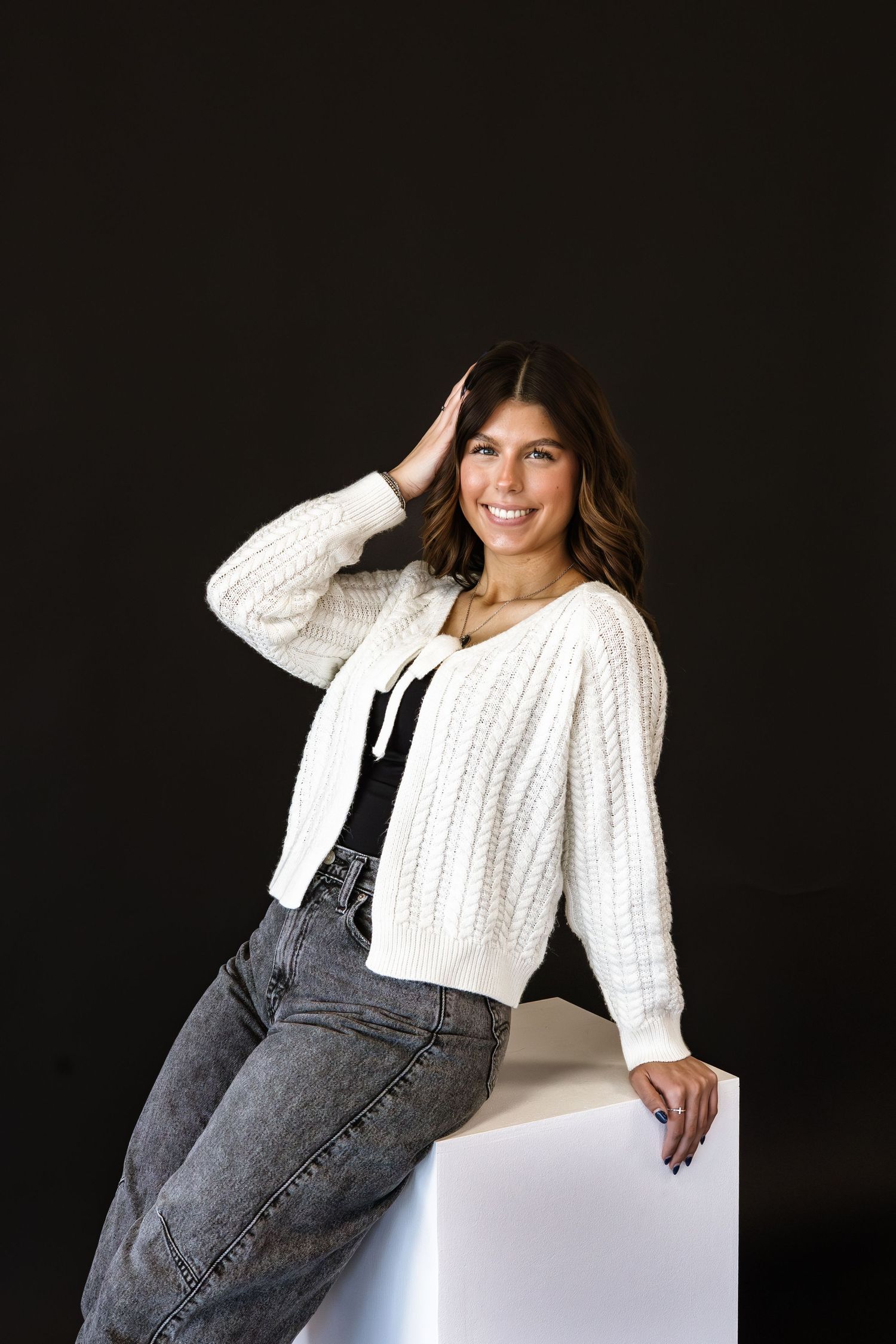 Smiling woman in white knit cardigan and gray jeans leaning on white cube against dark background.