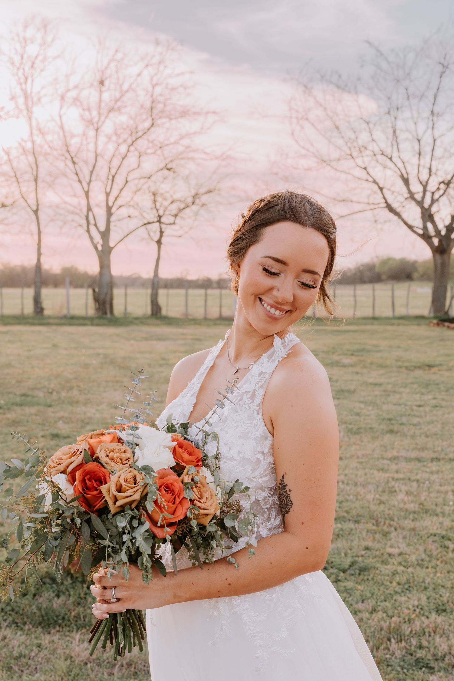 Smiling bride in white halter gown holds orange rose bouquet at golden hour in a grassy field with bare trees.