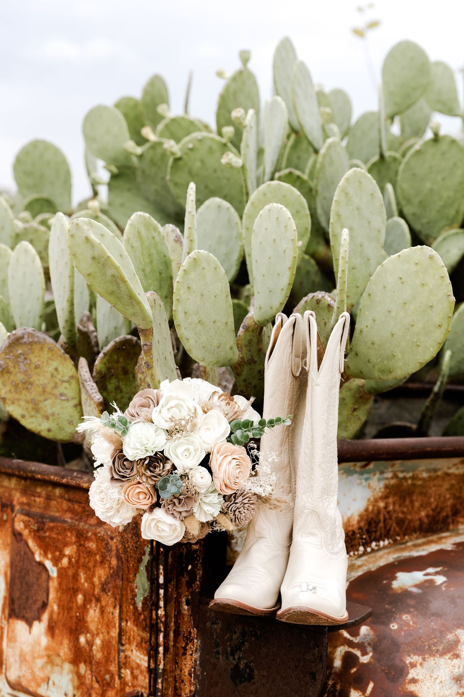 White cowboy boots and a neutral floral bouquet rest against a rustic metal container with prickly pear cactus backdrop.