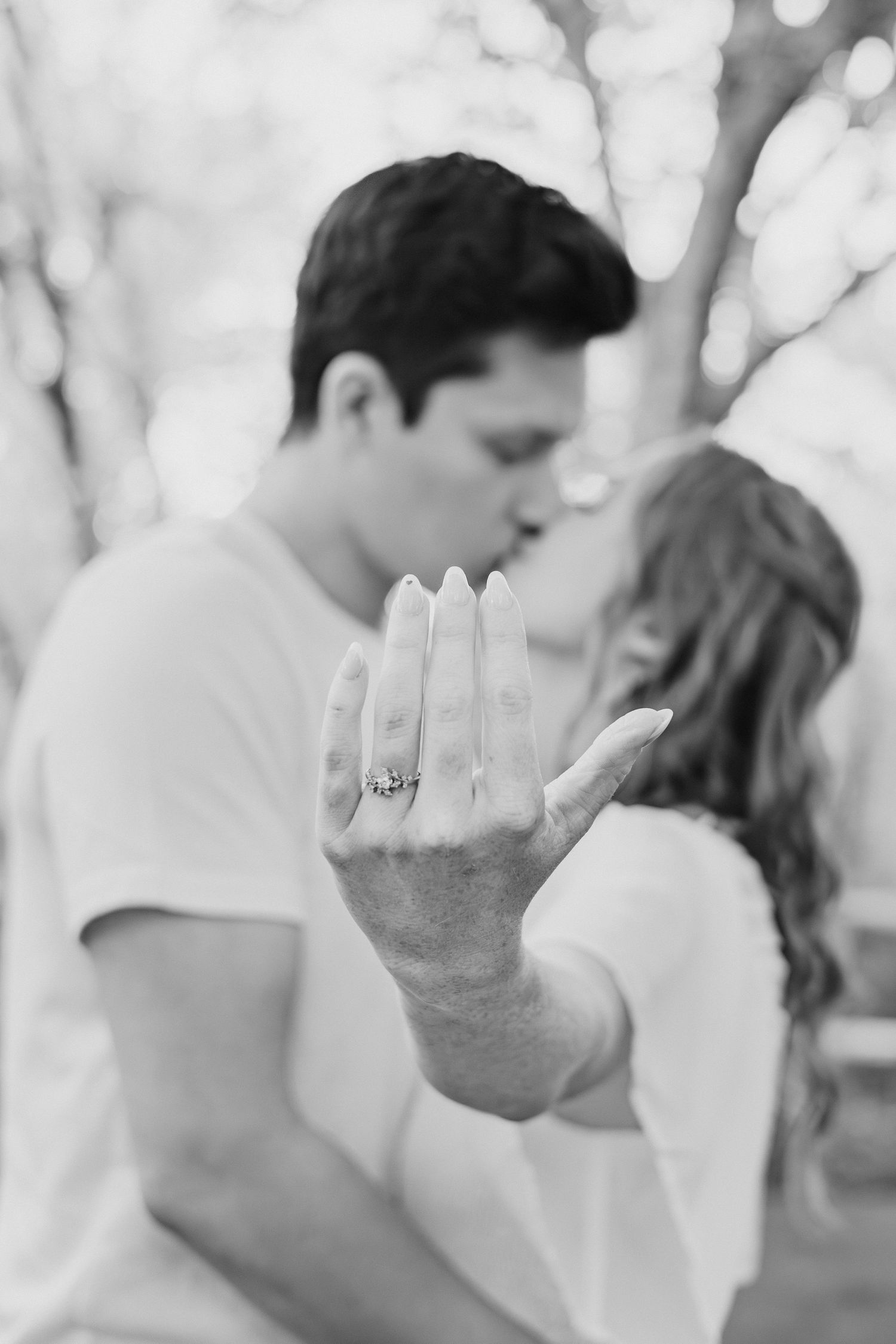 Black and white engagement photo of couple kissing while showing off diamond ring in foreground.