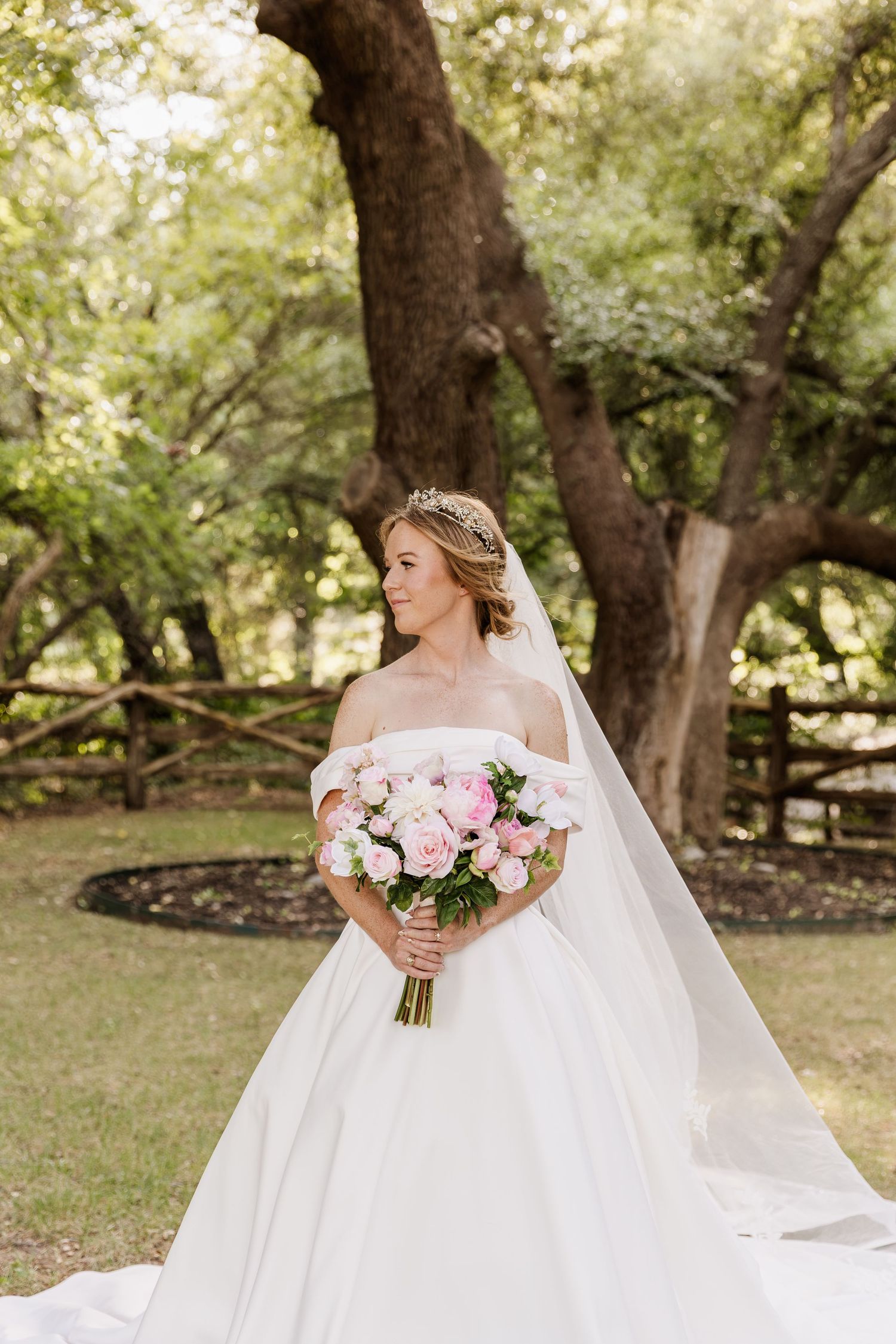 Bride in off-shoulder white ball gown holding pink bouquet, wearing tiara and veil, posing outdoors near large oak tree.