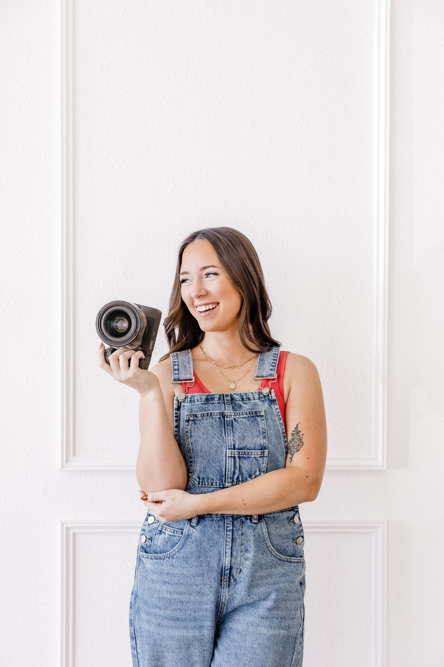 Smiling woman in denim overalls holding a camera lens against a white wall background.