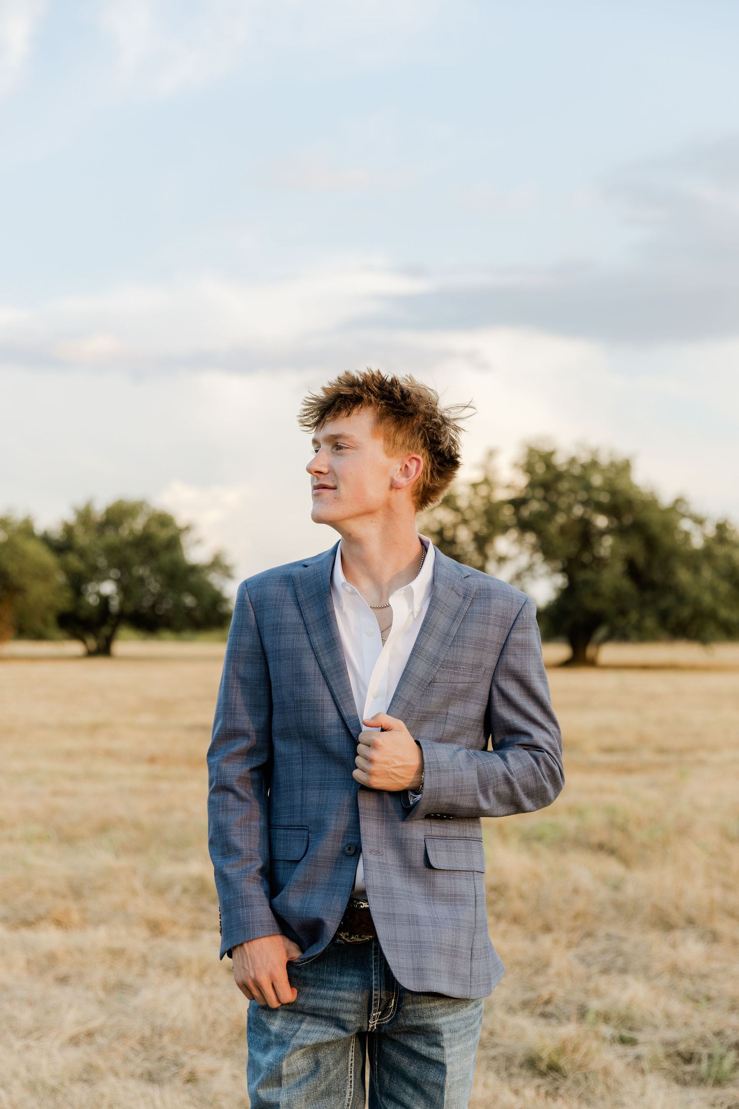 Young man in plaid blazer and jeans standing in a golden field with trees and cloudy sky backdrop.
