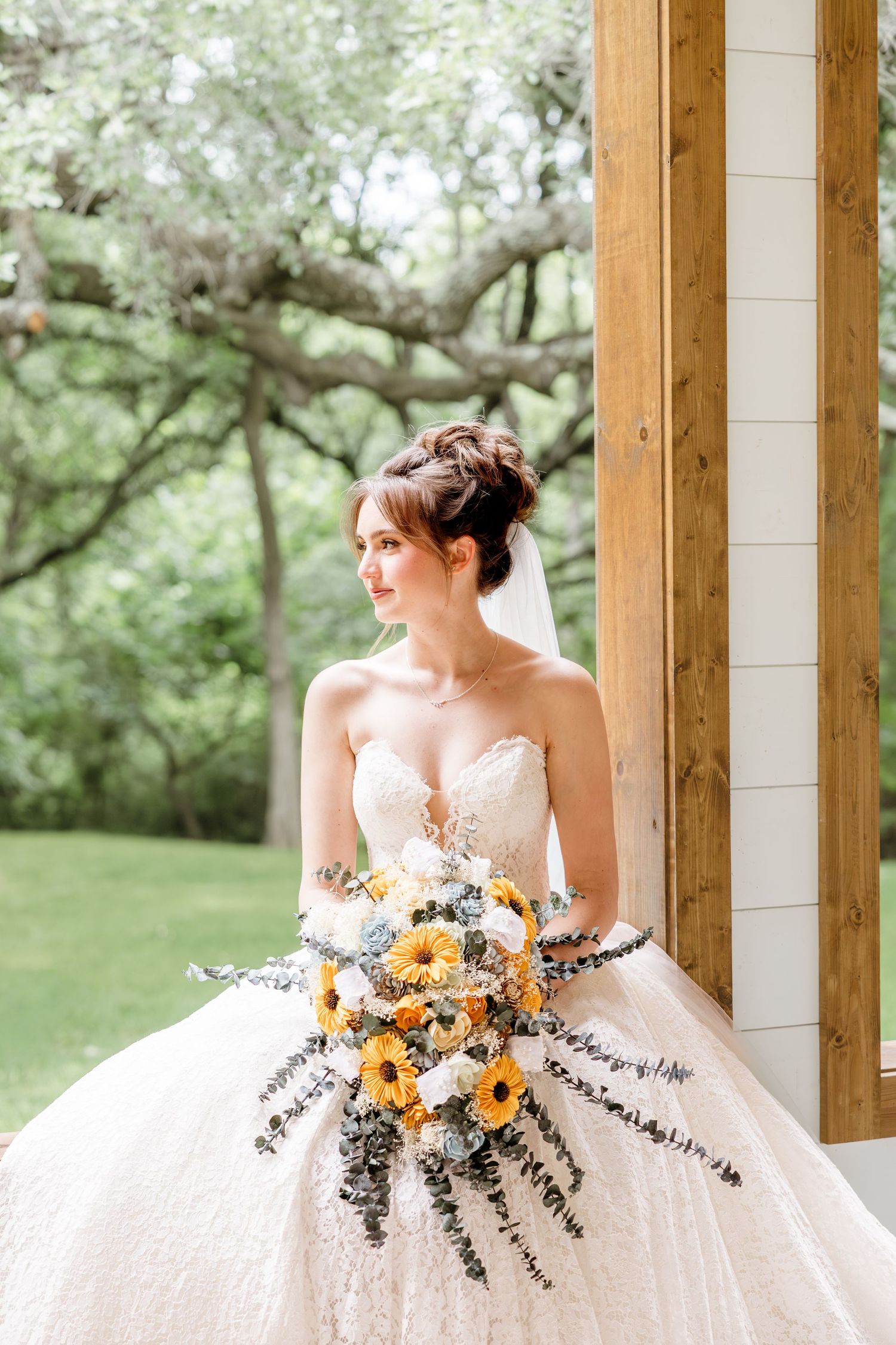 Bride in strapless lace ball gown holding sunflower bouquet, posing by wooden pillar with lush green trees background.