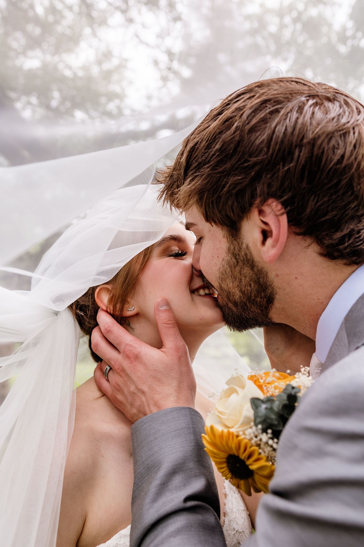 Bride and groom share a romantic kiss under a flowing veil, with sunflowers and greenery in the background.