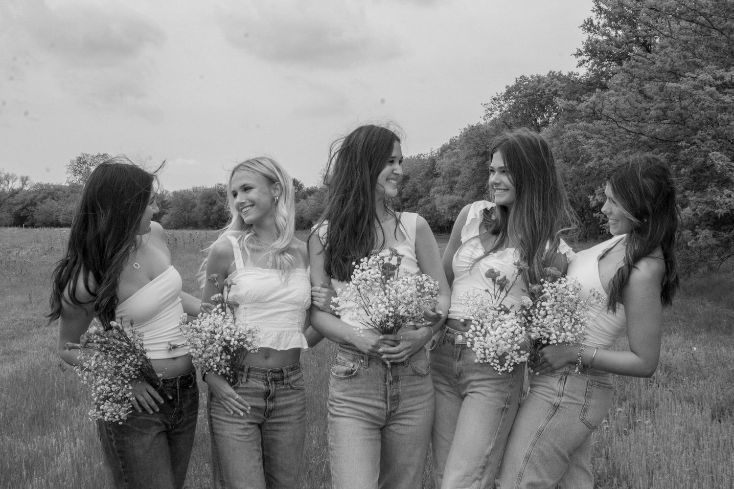 A black and white photograph of young people in casual denim outfits holding baby's breath bouquets in a grassy field.
