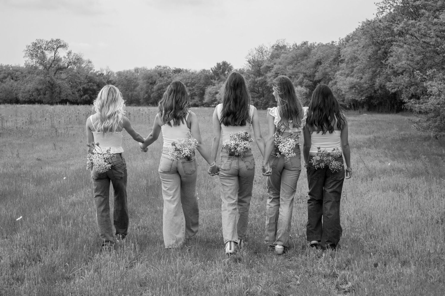 Five friends in denim jeans hold hands while walking away in a grassy field.