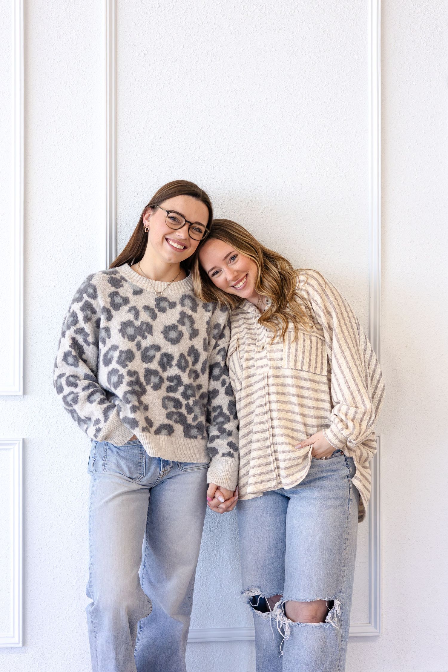 Two people wearing cozy sweaters and jeans pose casually against a white wall in matching casual outfits.