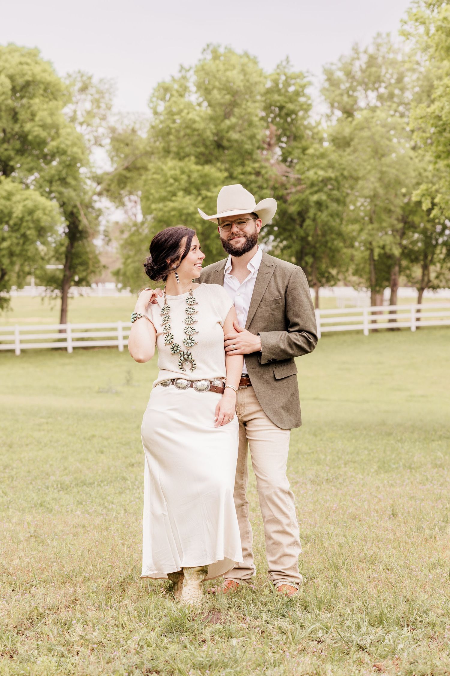 Couple in western attire standing in green field, man in cowboy hat and blazer, woman in white dress with boots.