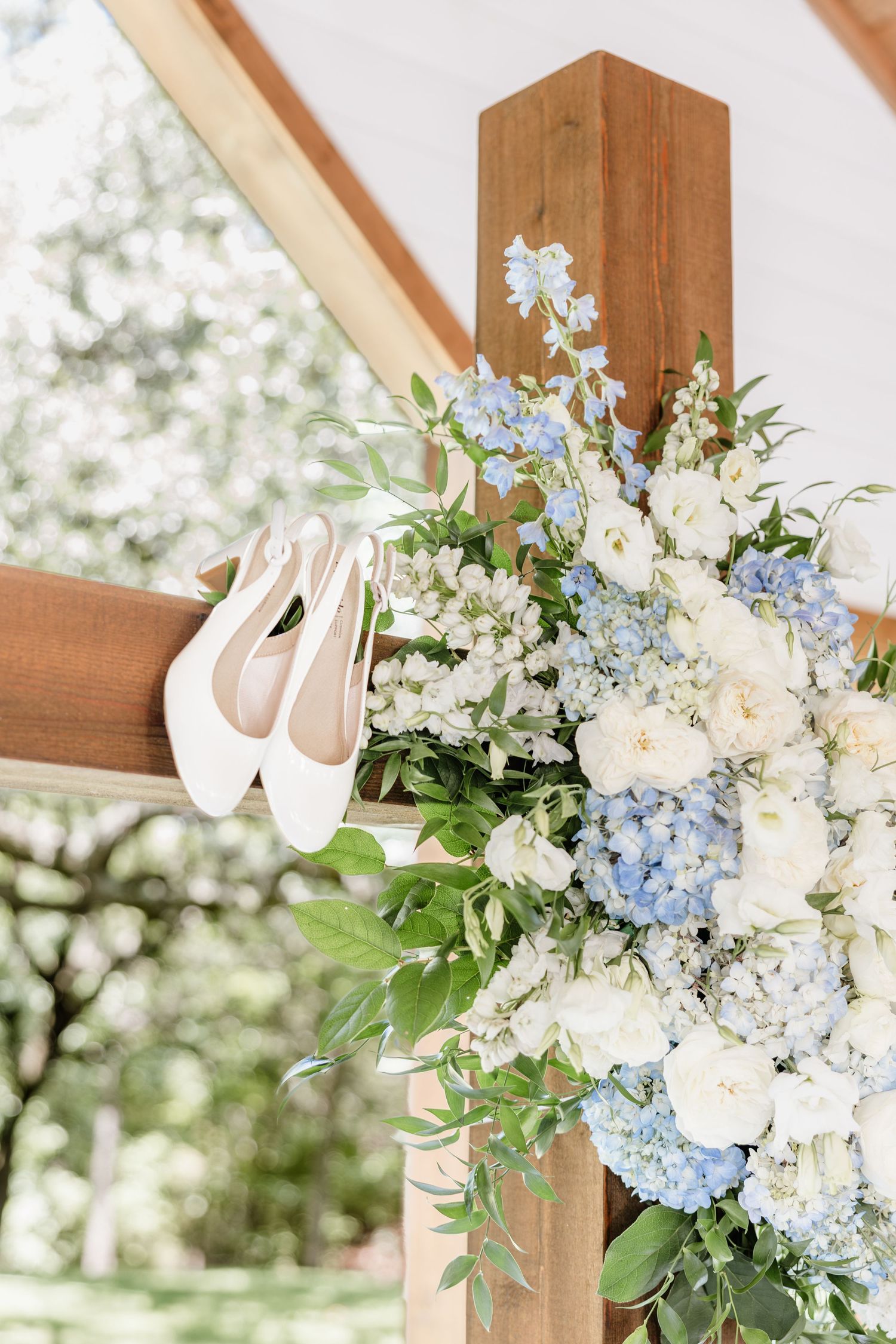 White bridal heels hang on a wooden arch adorned with blue hydrangeas and white roses at an outdoor wedding ceremony.