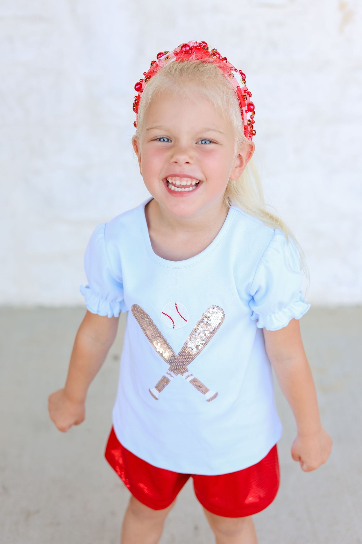 Young child wearing blue shirt with baseball bats and red shorts with flower crown.