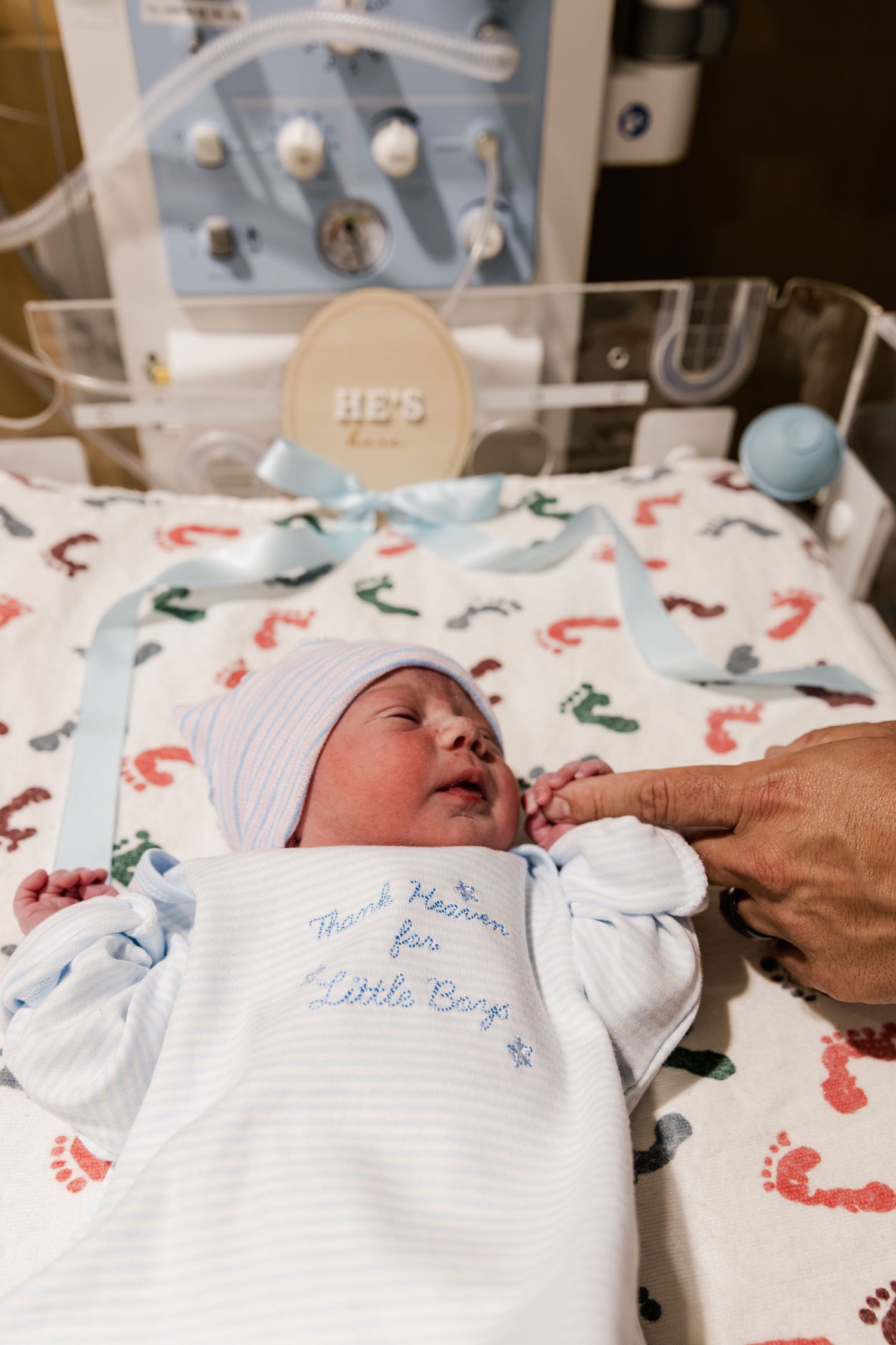 Newborn baby boy in hospital bassinet wearing white gown reading Thank Heaven for Little Boys with blue hat.