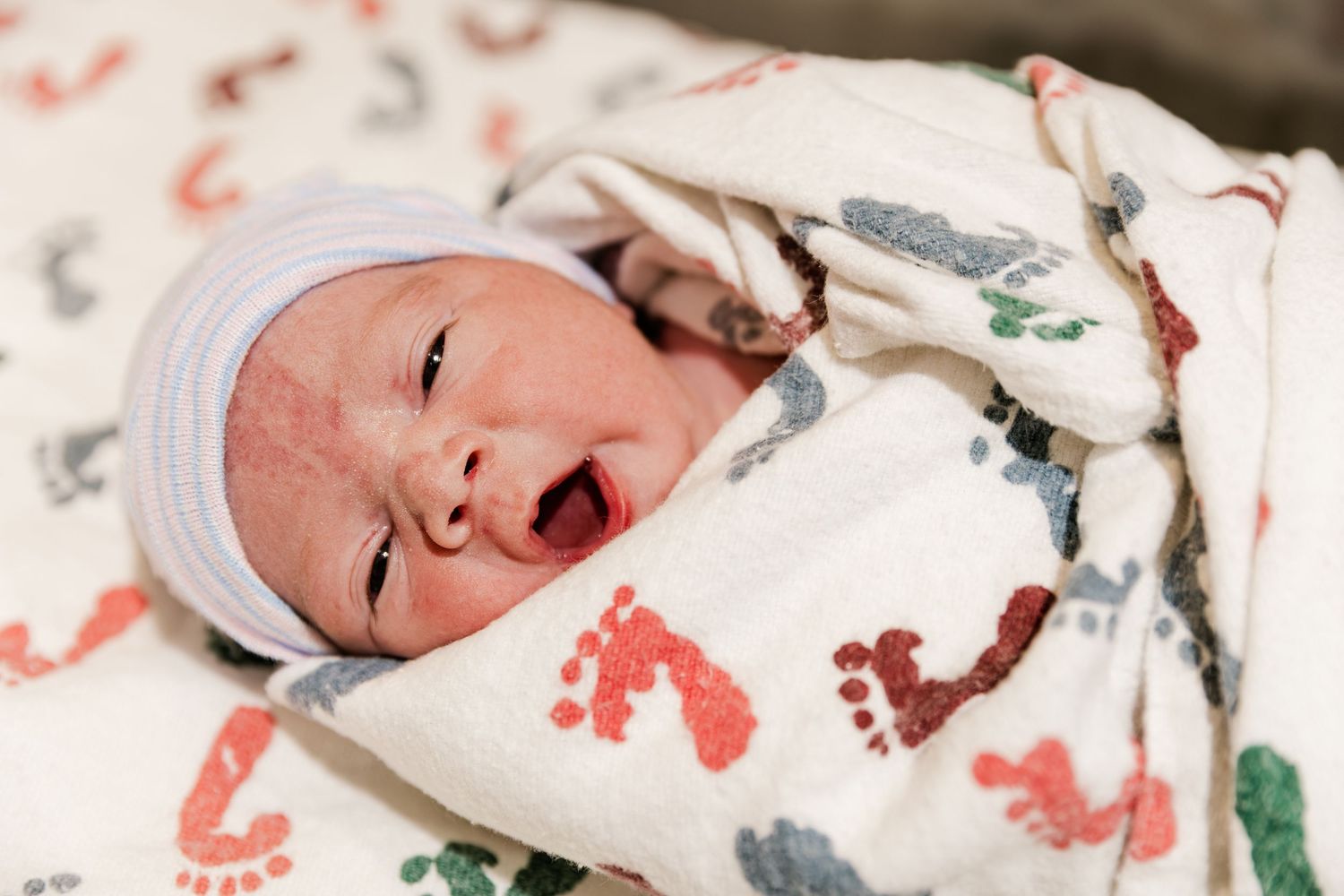 Newborn baby wrapped in a colorful footprint swaddle blanket wearing a white striped hat, yawning with mouth open.
