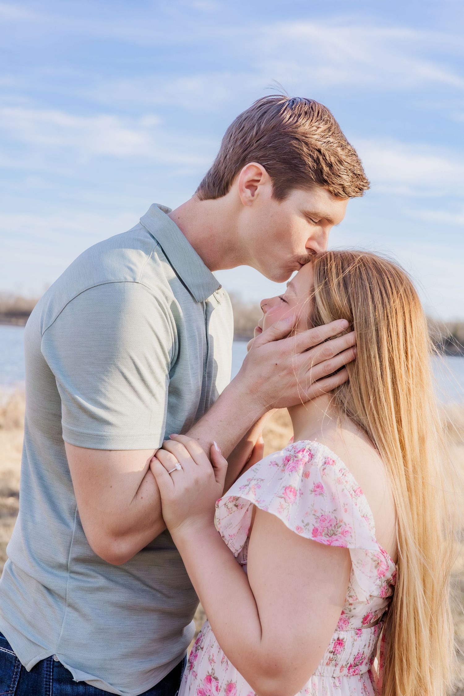 Romantic couple sharing a tender kiss outdoors near water, woman in floral dress, man in gray polo shirt.