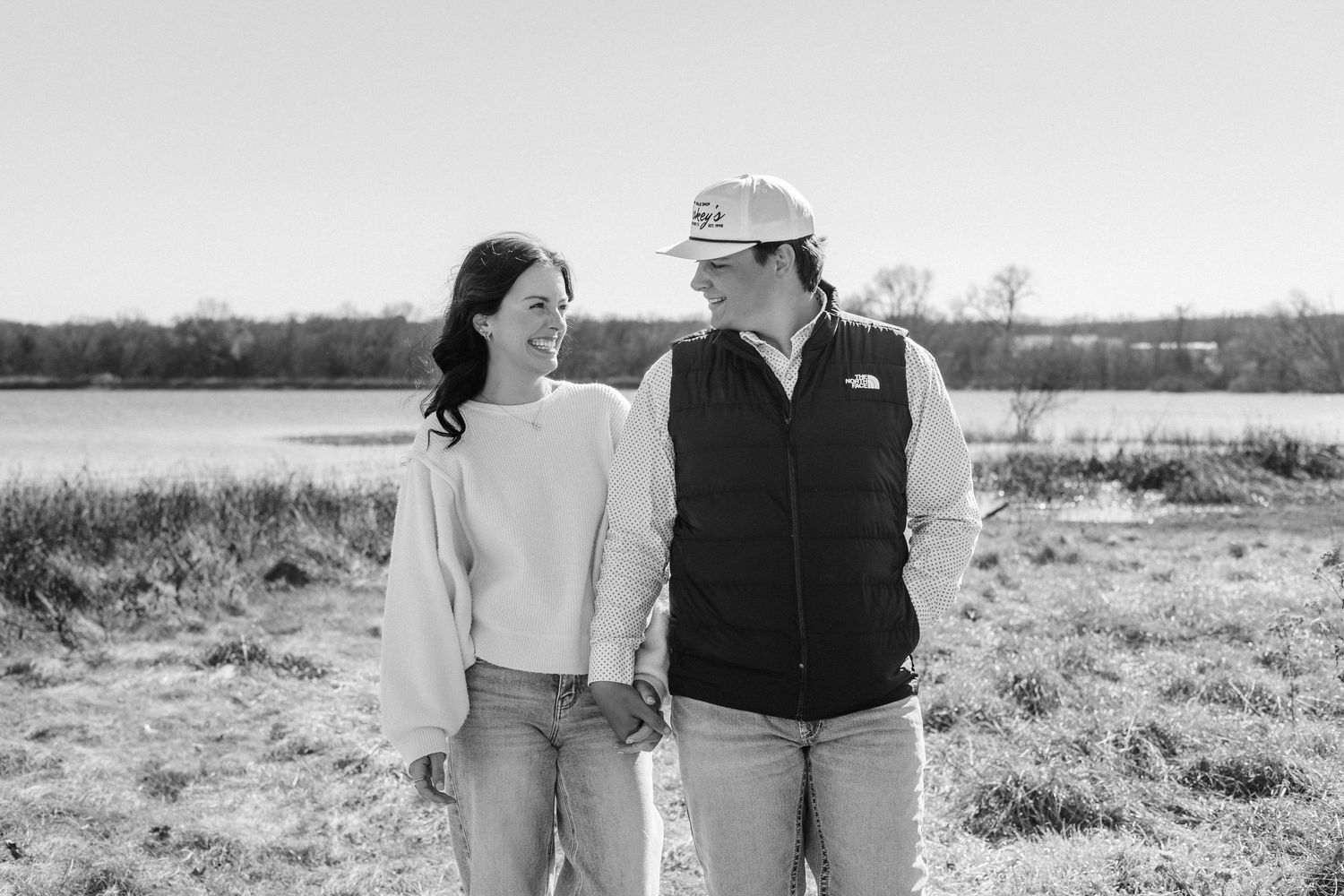 Couple smiling and holding hands outdoors near a lake on a sunny day in a black-and-white photo.