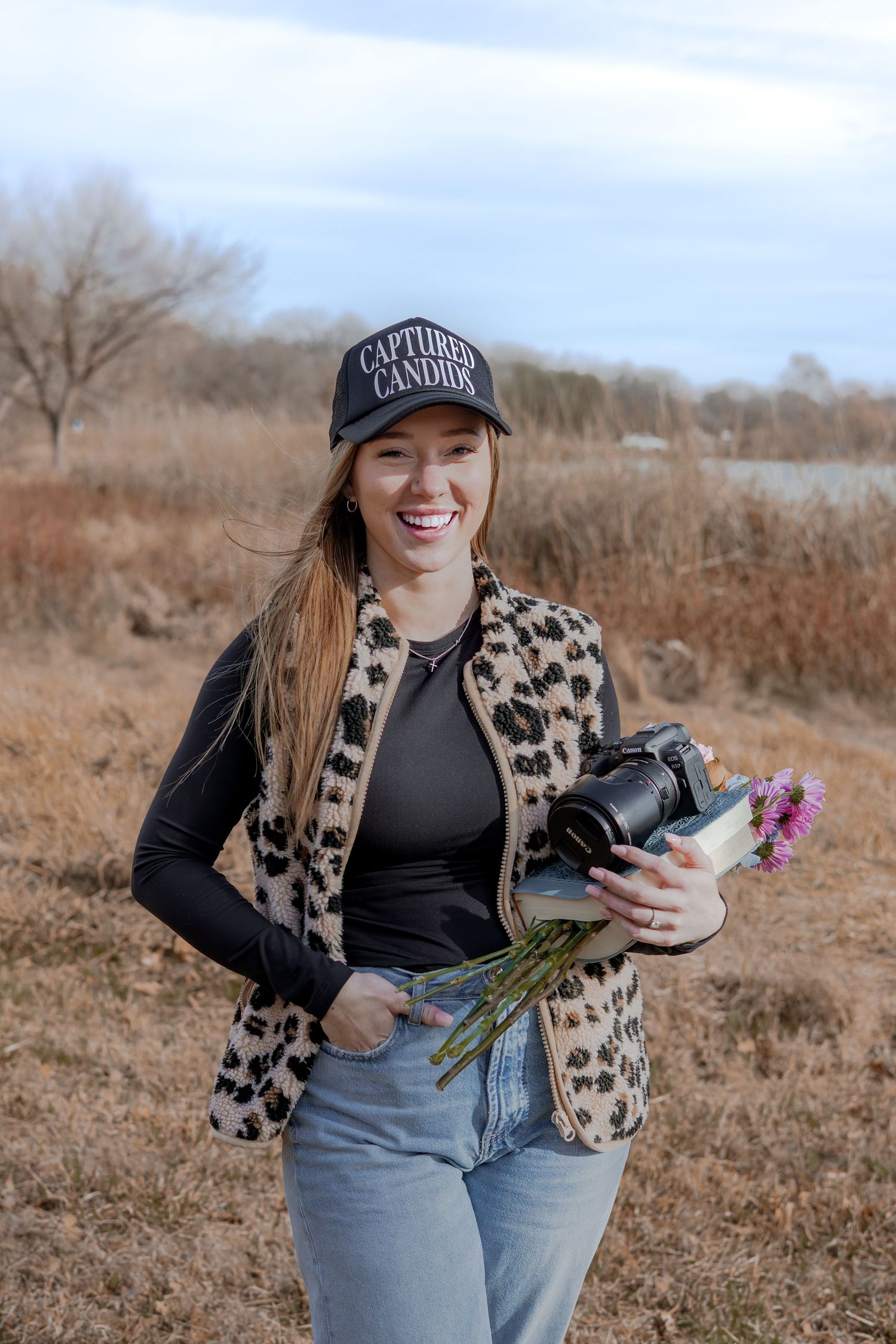 A photographer in a leopard print vest and baseball cap poses outdoors with camera equipment in a winter field.