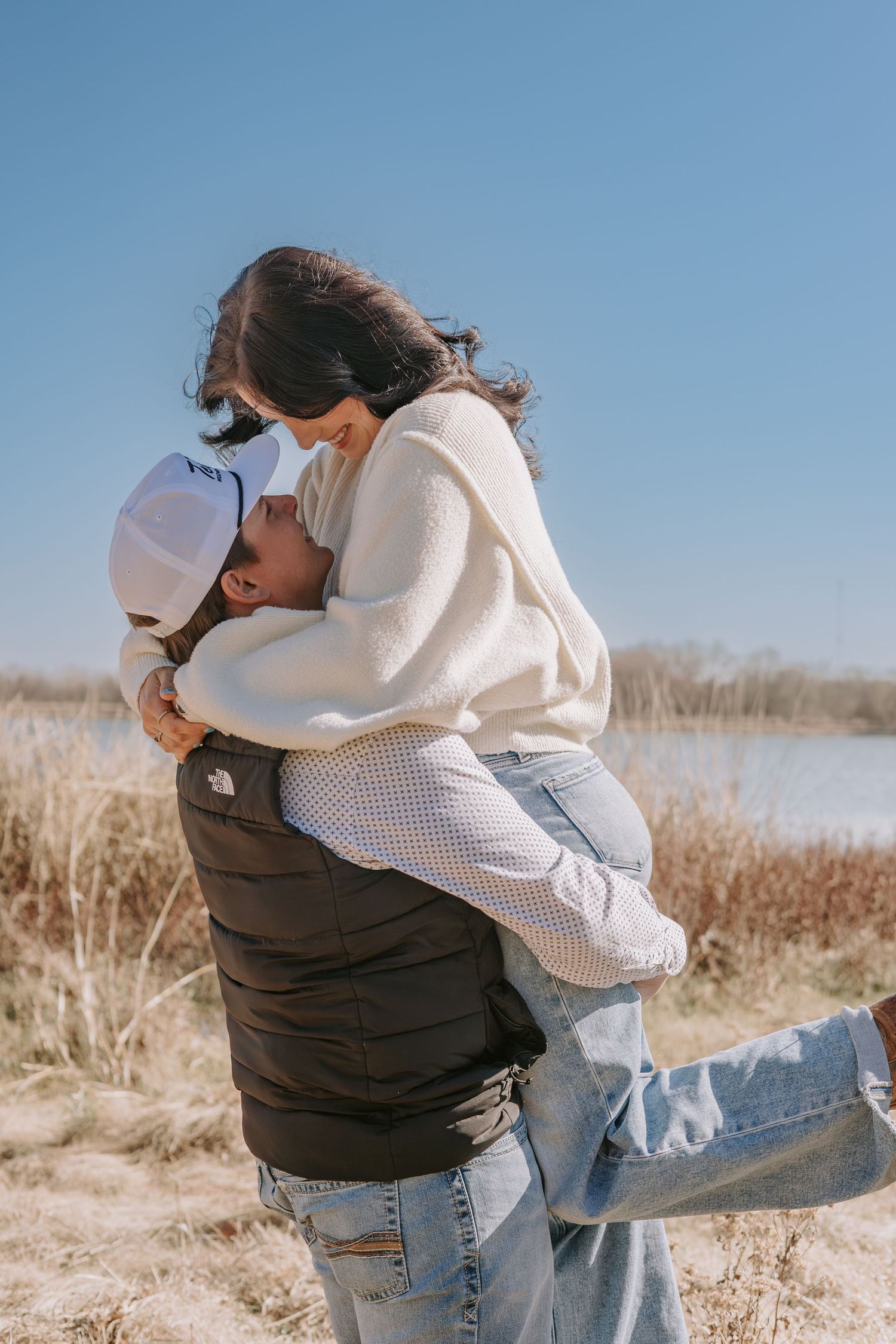 Couple embracing outdoors near a lake, woman lifted in man's arms, wearing casual winter clothes on a sunny day.