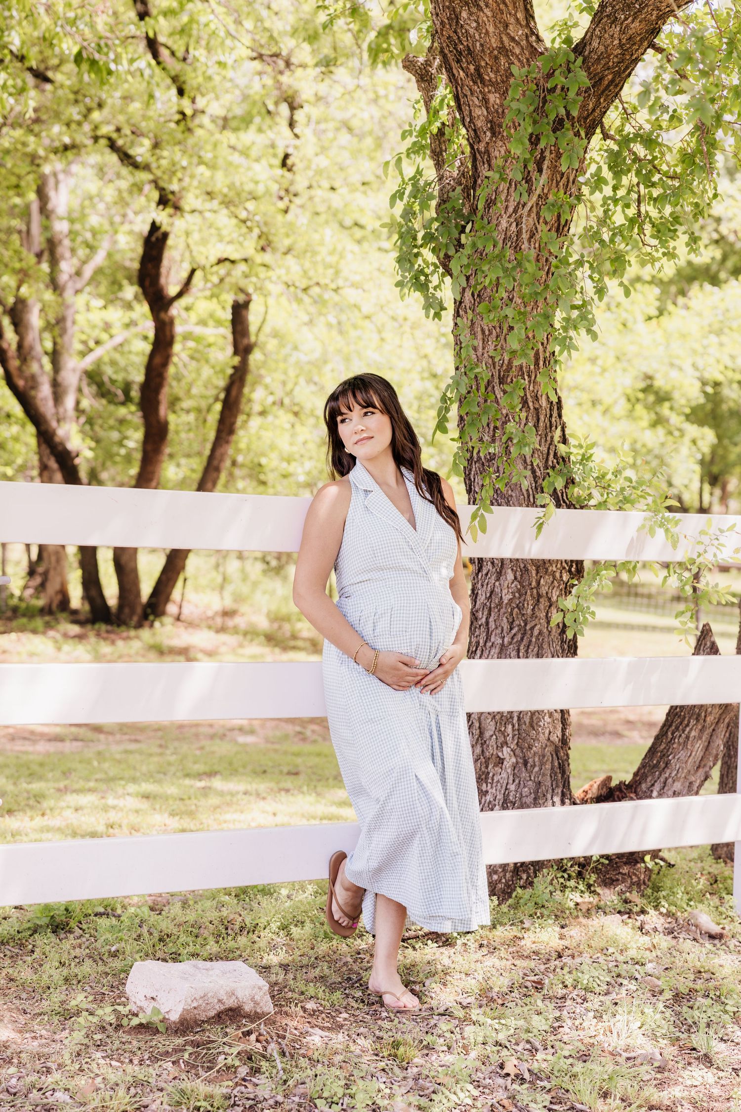 Pregnant woman in light blue dress holding belly, leaning against white fence surrounded by green trees.