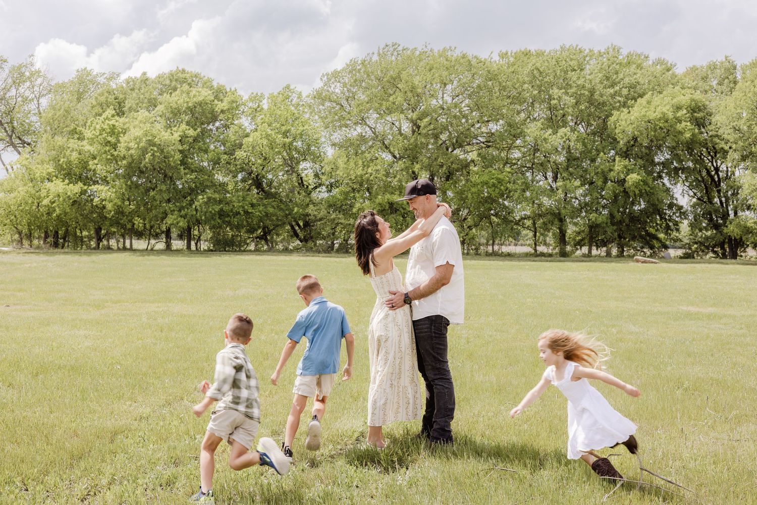 Family with three young children playing in a lush green park on a cloudy day, parents embracing while kids run around.