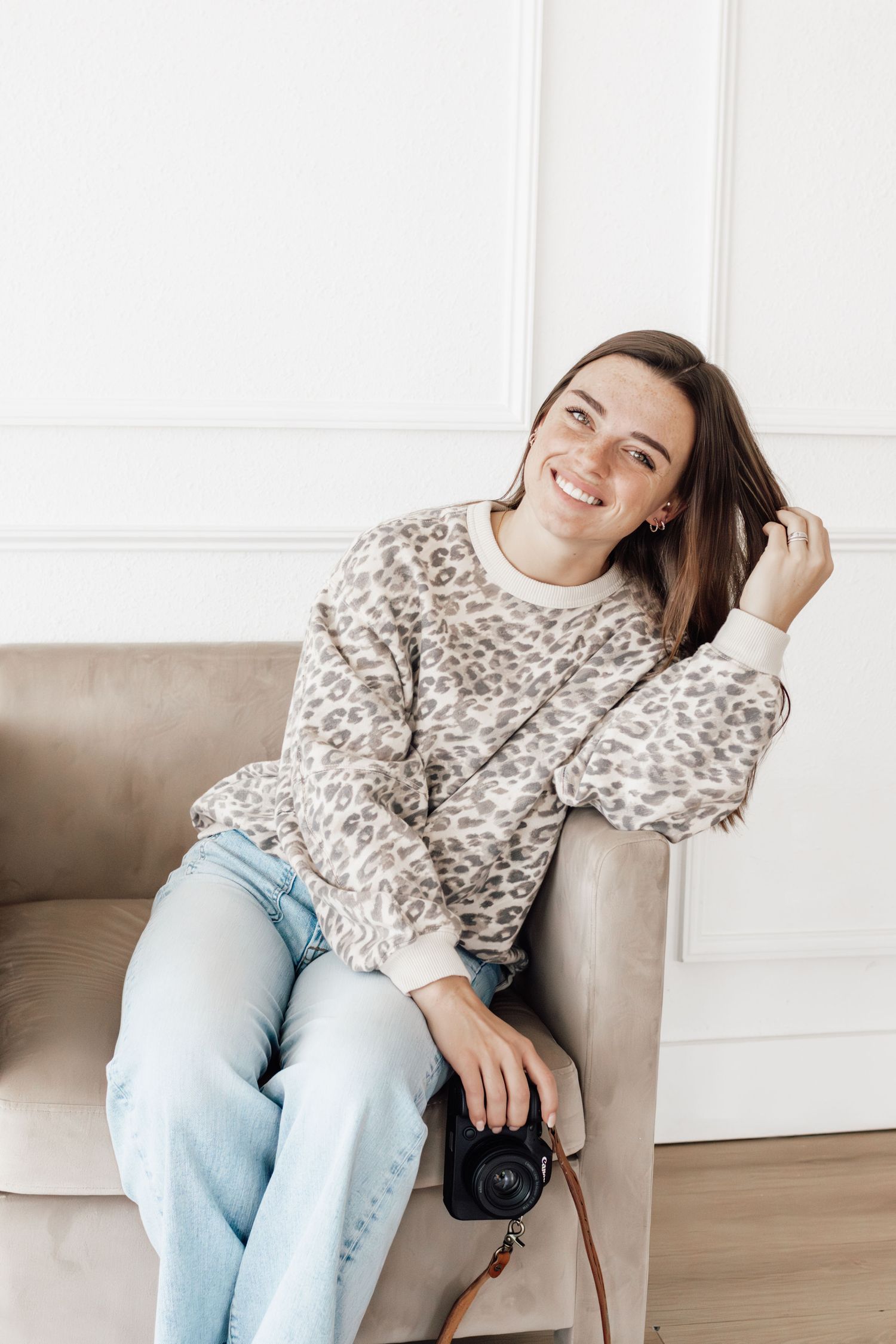 Casual lifestyle portrait of someone in a leopard print sweater and jeans relaxing on a beige couch with a camera.