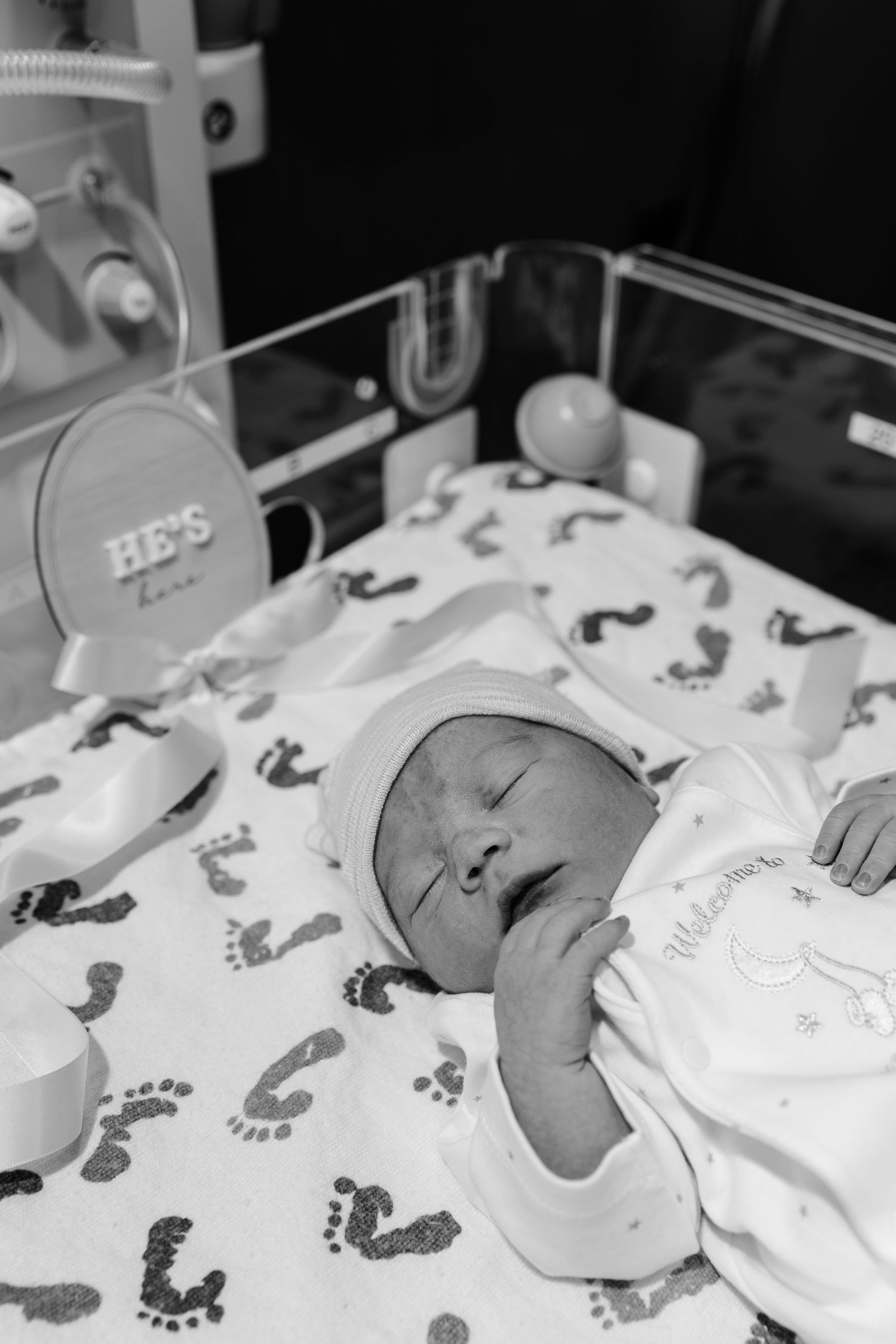 Black and white photo of a sleeping newborn baby in hospital bassinet with footprint blanket and He's Here sign.