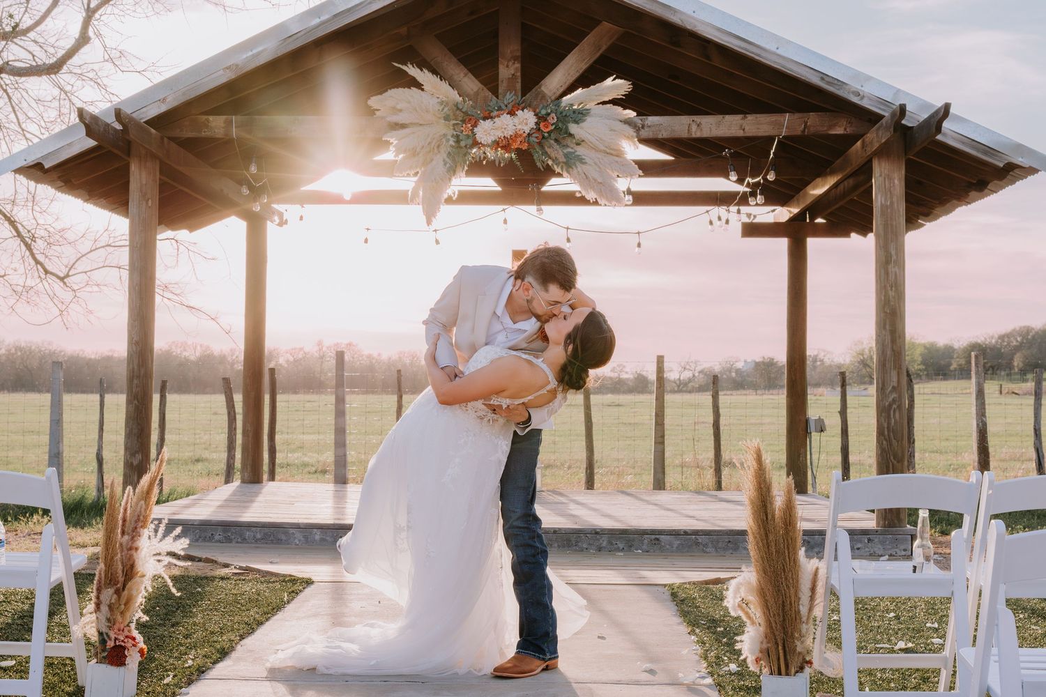 Bride and groom share a romantic kiss under a rustic wooden gazebo at sunset on a scenic farm wedding venue.