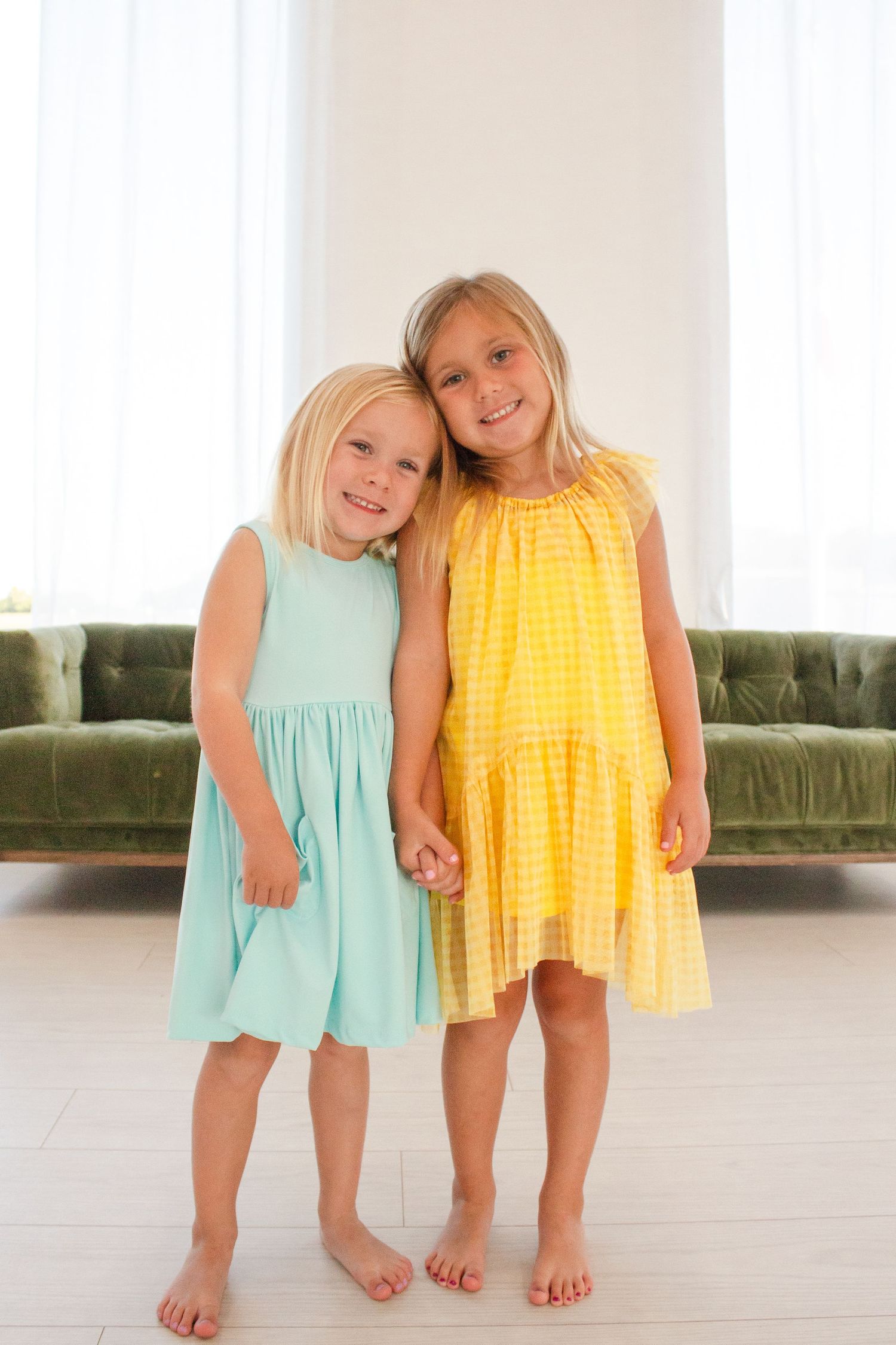 Two young children wearing summer dresses in mint green and yellow gingham stand together holding hands.