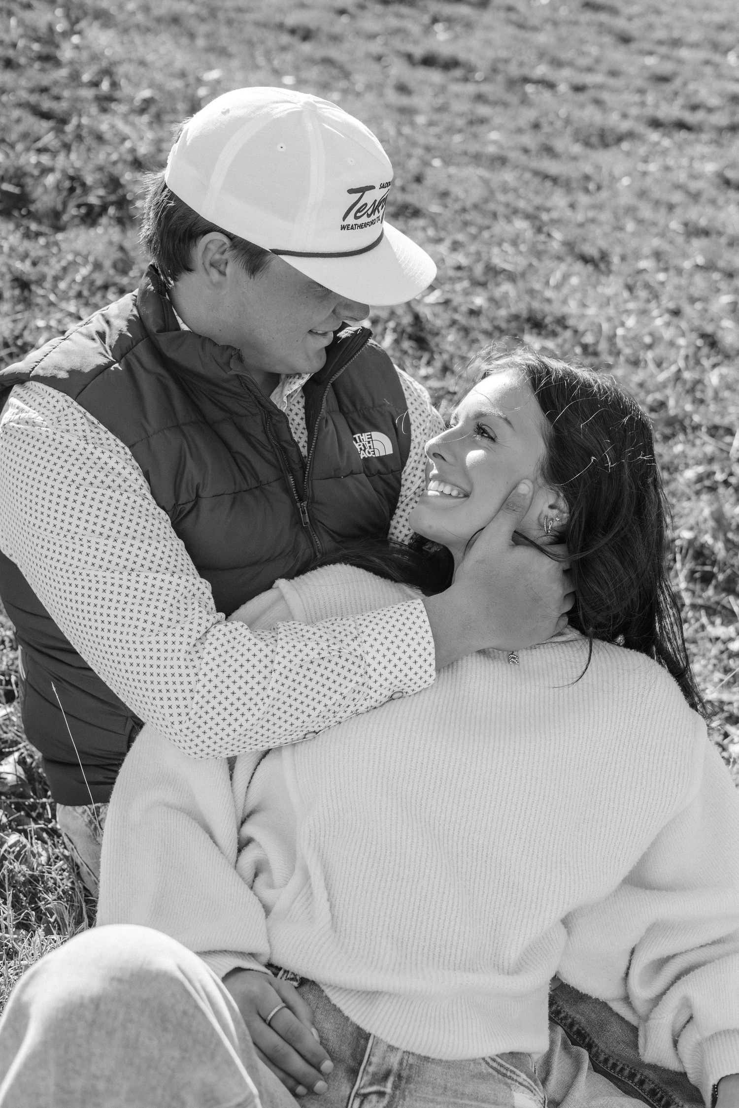 Black and white photo of a couple sitting in a field, smiling at each other in a tender romantic moment.