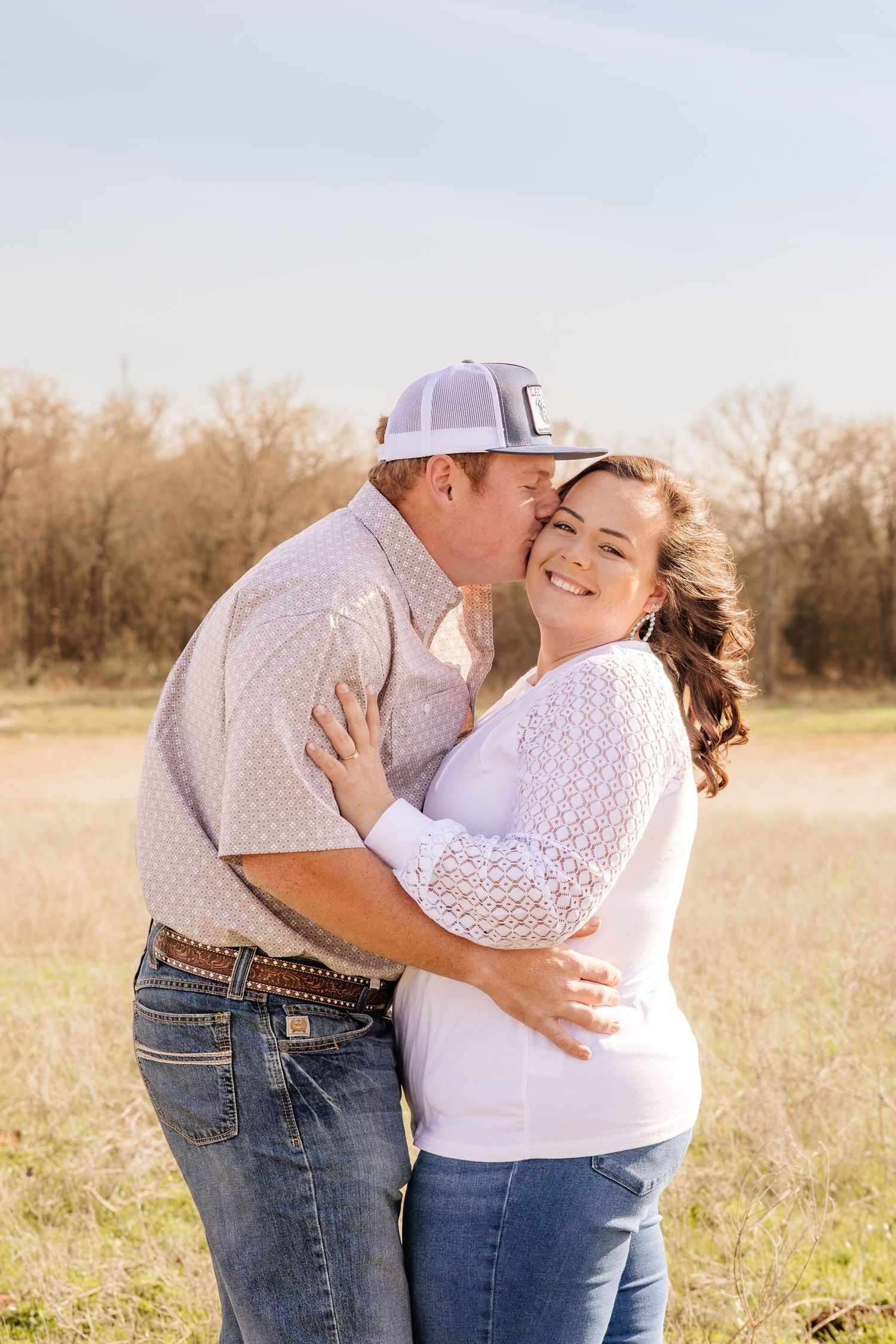 A couple embraces outdoors in a sunny field, man in cap and jeans kisses woman in white lace top smiling at camera.