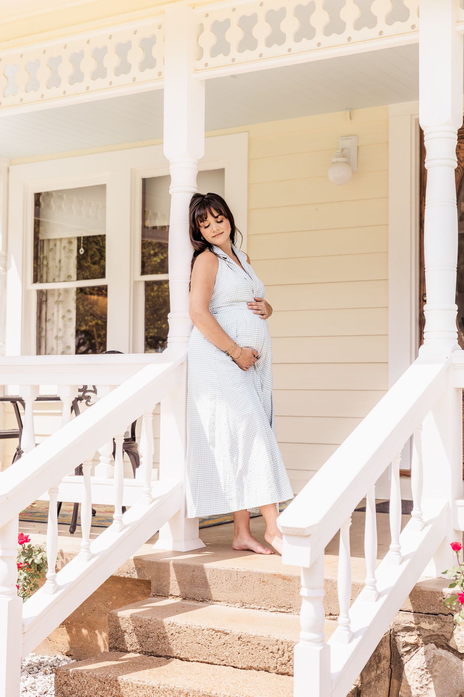 Pregnant woman in light blue dress standing on white porch steps, cradling baby bump in sunny outdoor setting.