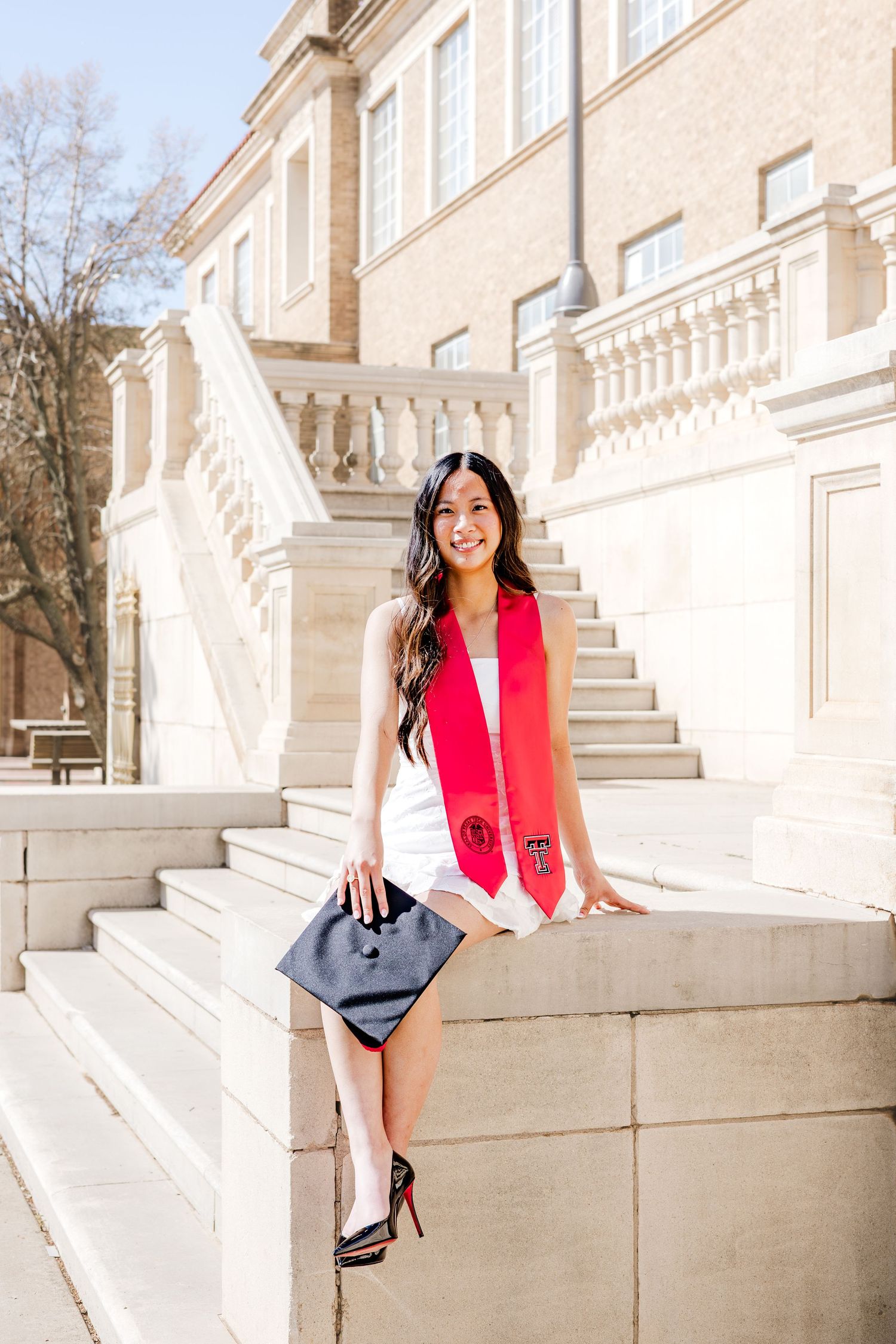 Smiling graduate in white dress and red stole holds cap on university steps.
