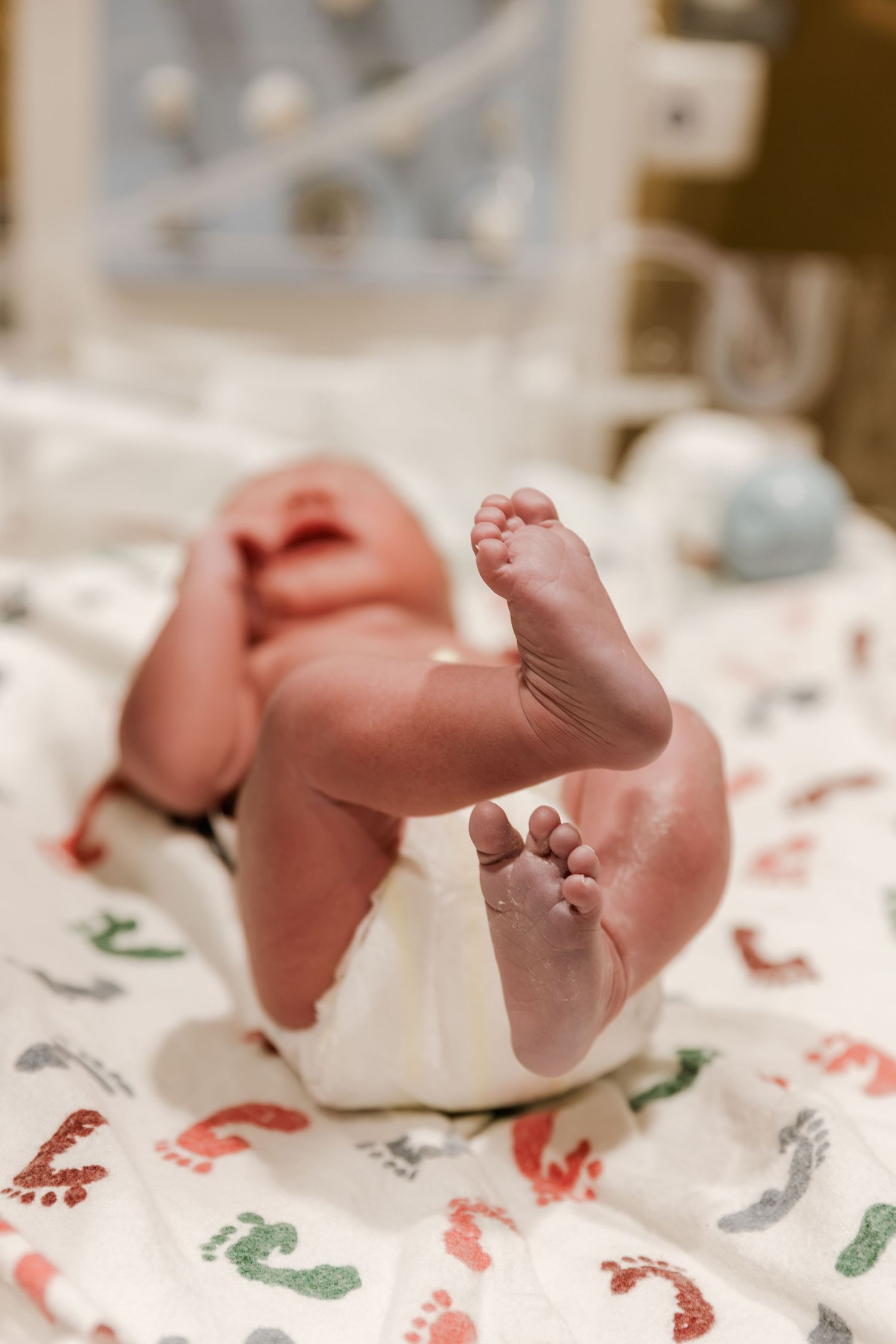 Newborn baby lying on a festive blanket with tiny feet raised in the air in a hospital nursery.